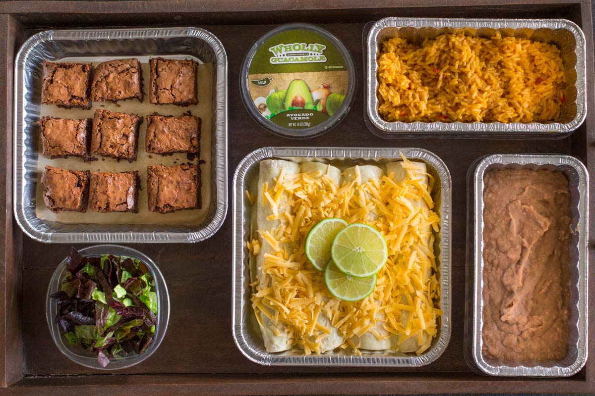 A wood tray with a pan of Honey Lime Chicken Enchiladas before being cooked, a pan of Healthy Crockpot Refried Beans, a pan of Spanish Rice, a container of Wholly Guacamole, a side salad, and a pan of Extra Thick and Fudgy Homemade Brownies. 