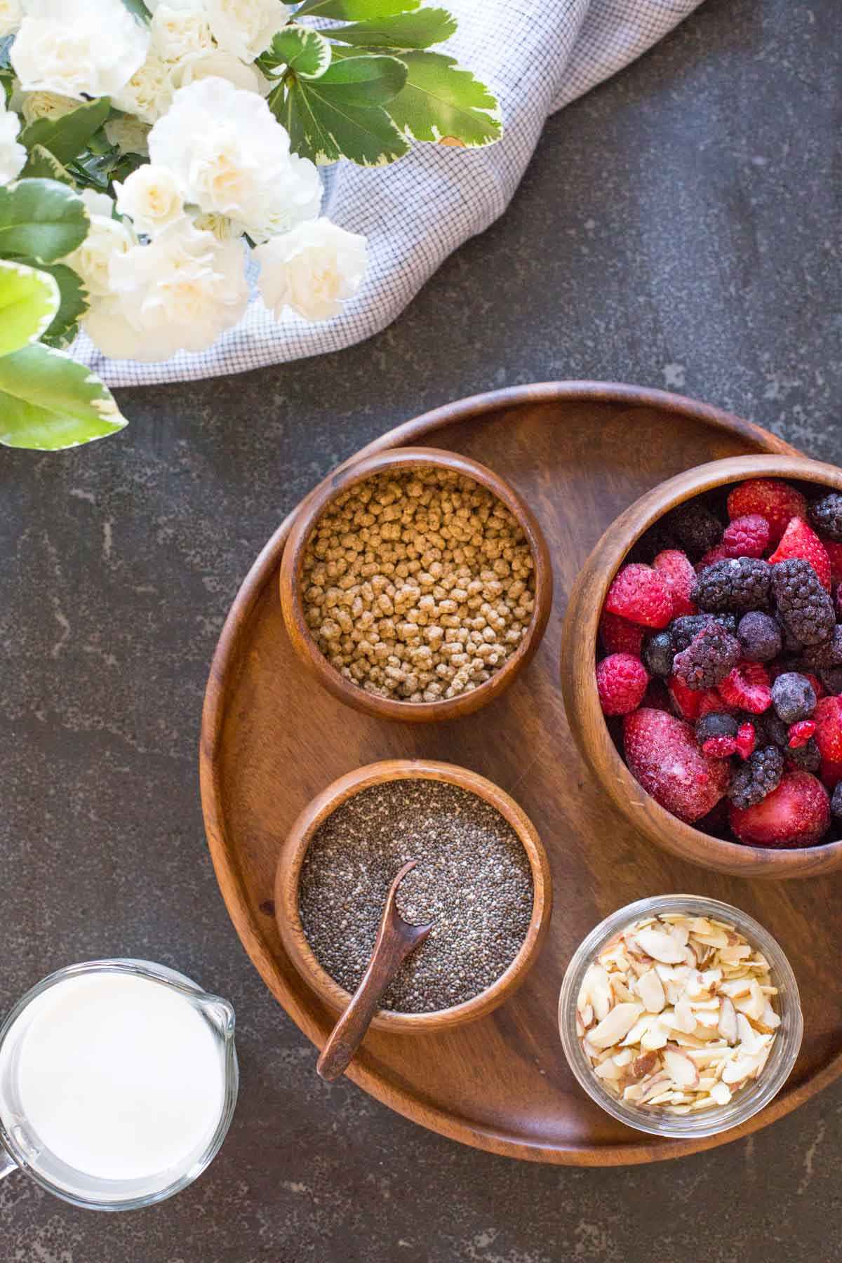 All the ingredients for the Chia Pudding Parfaits - A wood plate with a small wood bowl of bran nuggets, a small wood bowl of chia seeds, a small glass dish of sliced almonds, and a wood bowl of frozen mixed berries, sitting next to a small pour cup of milk.