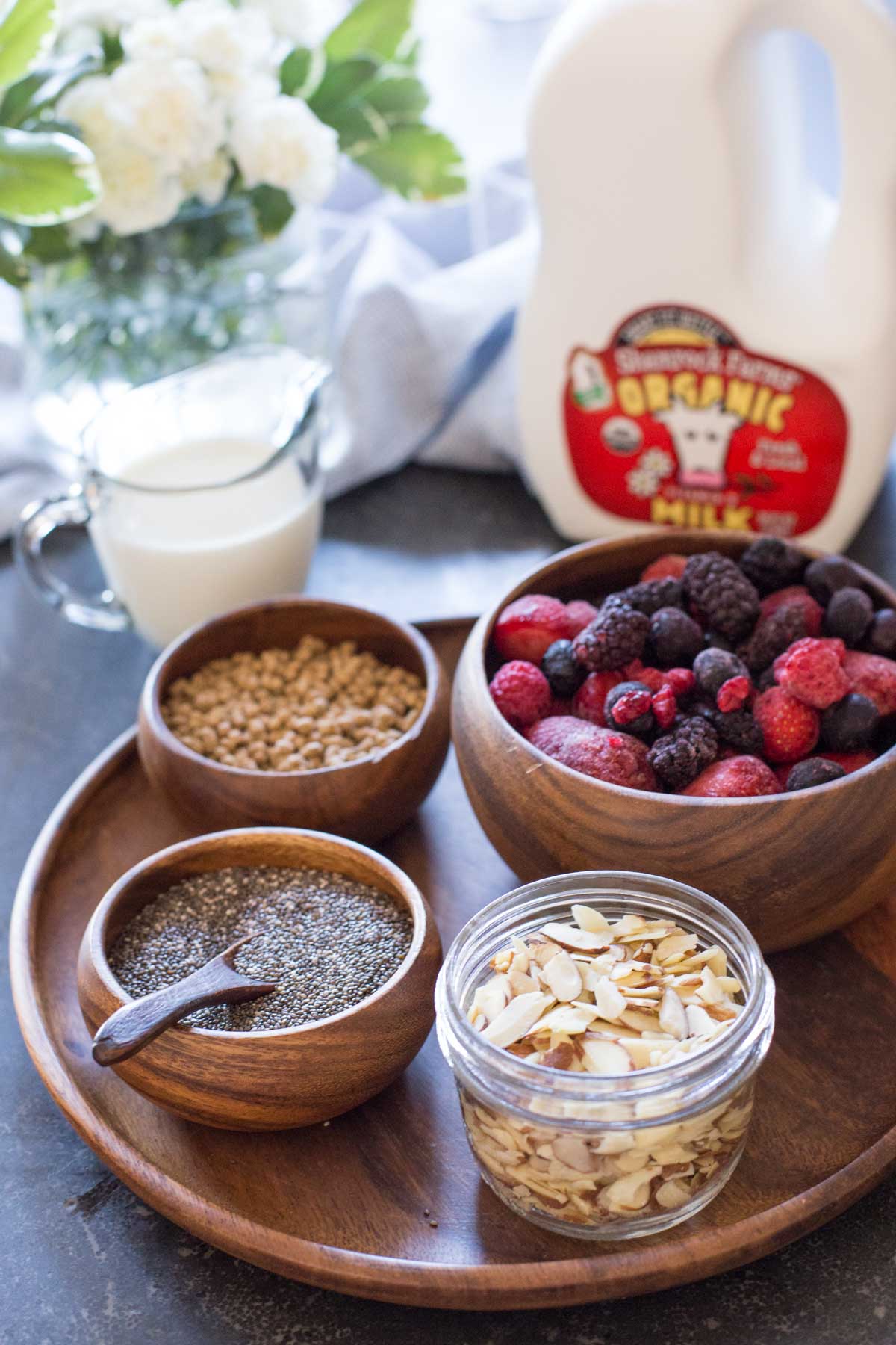 All the ingredients for the Chia Pudding Parfaits - A wood plate with a small wood bowl of bran nuggets, a small wood bowl of chia seeds, a small glass dish of sliced almonds, and a wood bowl of frozen mixed berries, sitting next to a milk jug and a small pour cup of milk. 