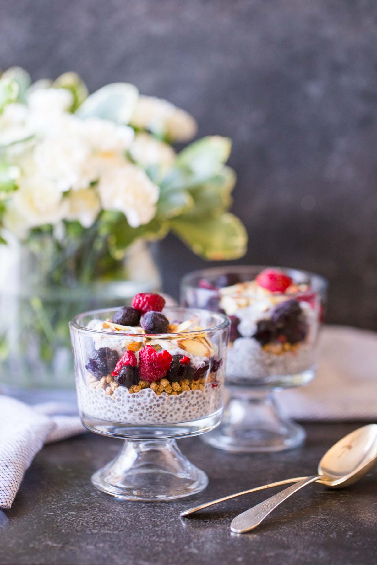 Chia Pudding Parfaits assembled in two small glass dishes, with two spoons next to them and a flower arrangement in the background. 