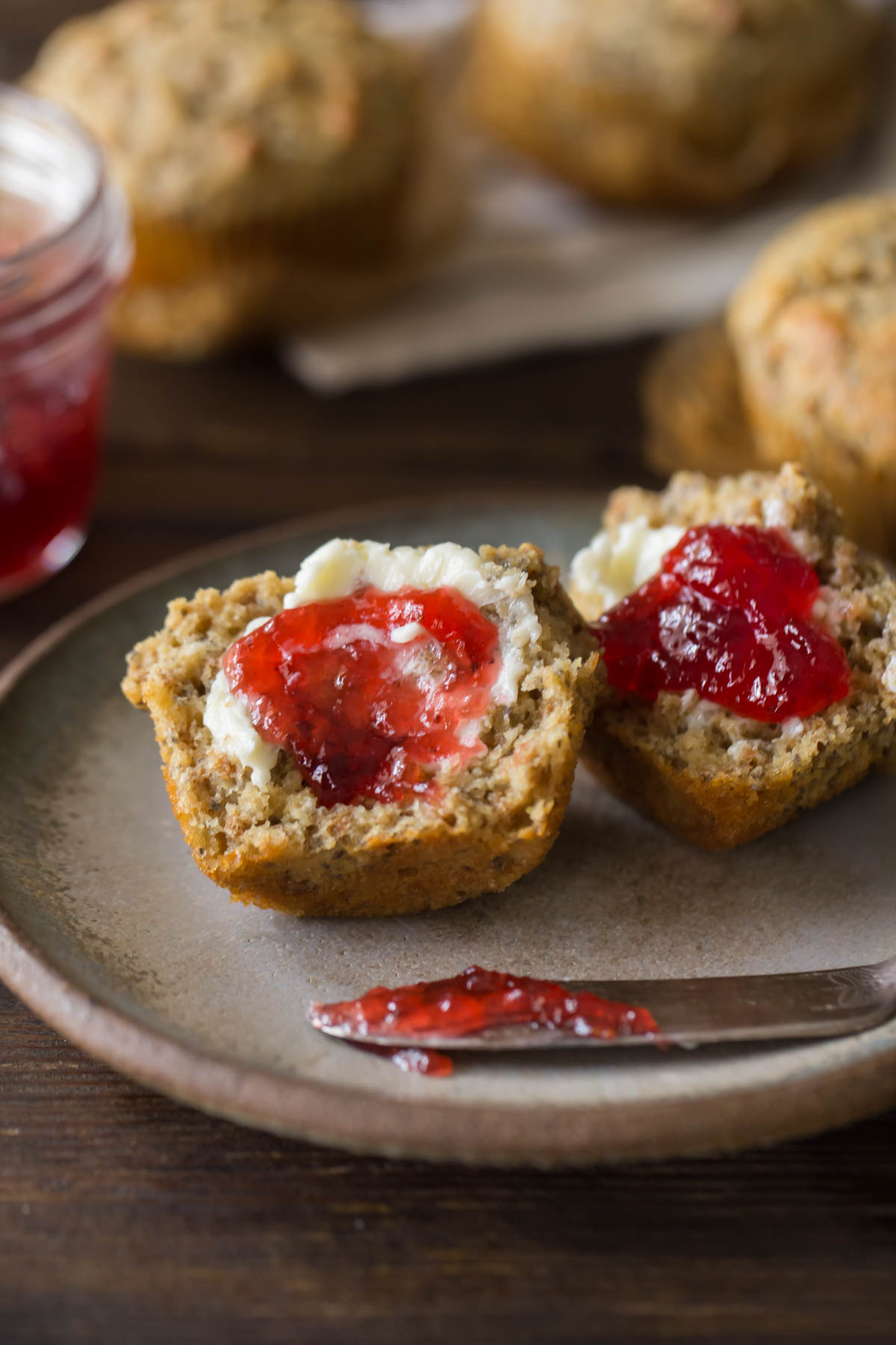 A Honey Bran Muffin cut in half with butter and jam on it, sitting on a plate with the jam knife, with three whole Honey Bran Muffins next to the plate along with the jar of jam.  