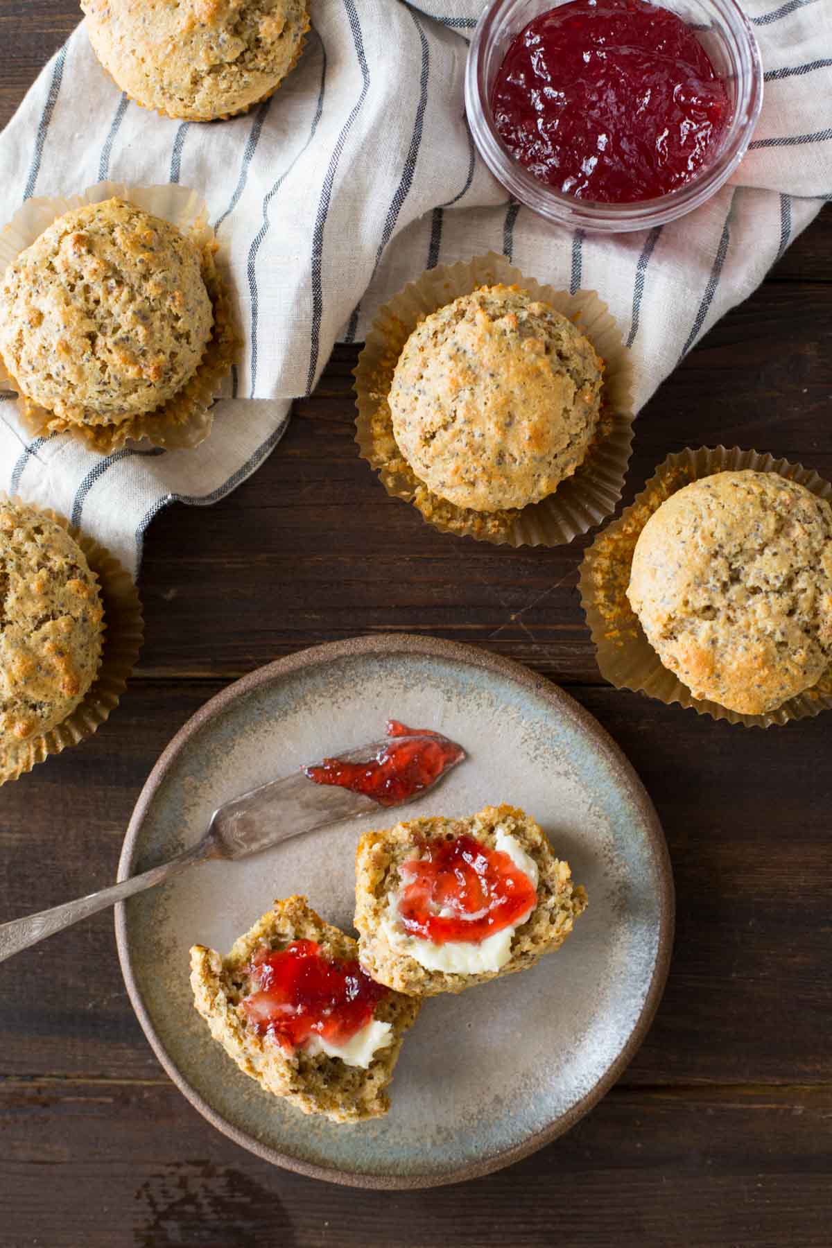 A Honey Bran Muffin cut in half with butter and jam on it, sitting on a plate with the jam knife, with five whole Honey Bran Muffins next to the plate along with the jar of jam.  