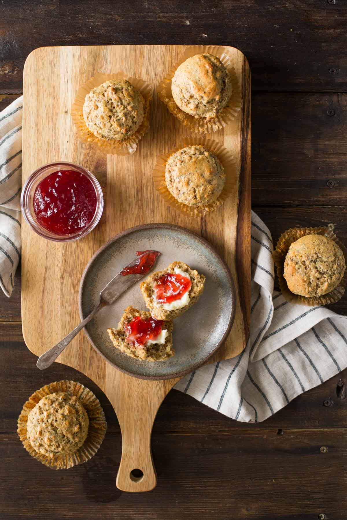 A Honey Bran Muffin cut in half with butter and jam on it, sitting on a plate with the jam knife, with five whole Honey Bran Muffins next to the plate along with the jar of jam.  