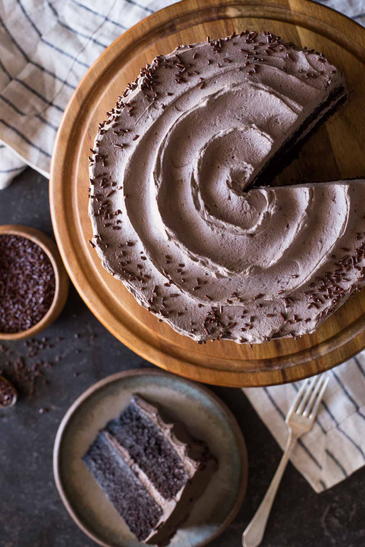 A slice of Dark Chocolate Cake With Whipped Cream Frosting on a plate, sitting next to the rest of the cake on a cake stand and a small wood bowl of chocolate sprinkles. 