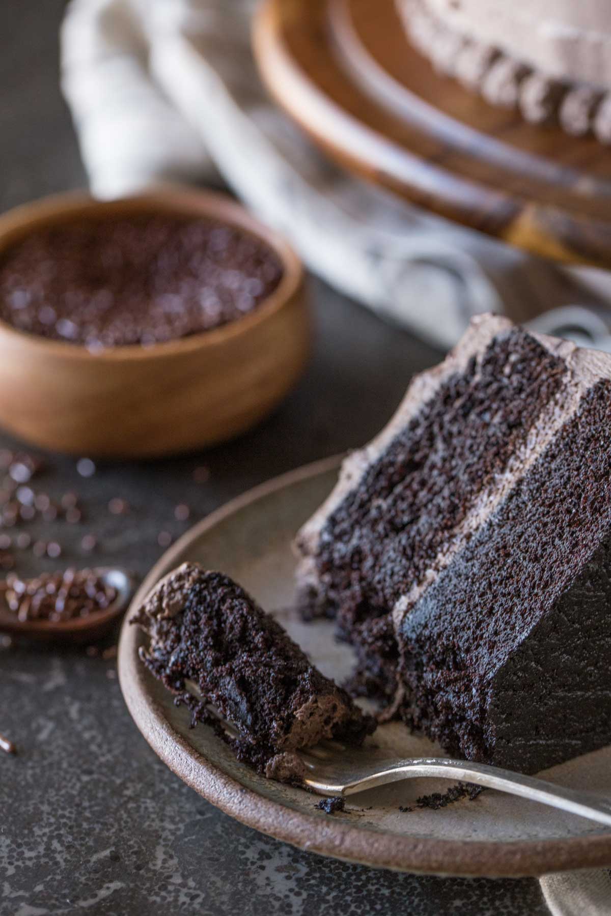 A slice of Dark Chocolate Cake With Whipped Cream Frosting on a plate along with a fork full of cake, and a small wood bowl of chocolate sprinkles in the background. 