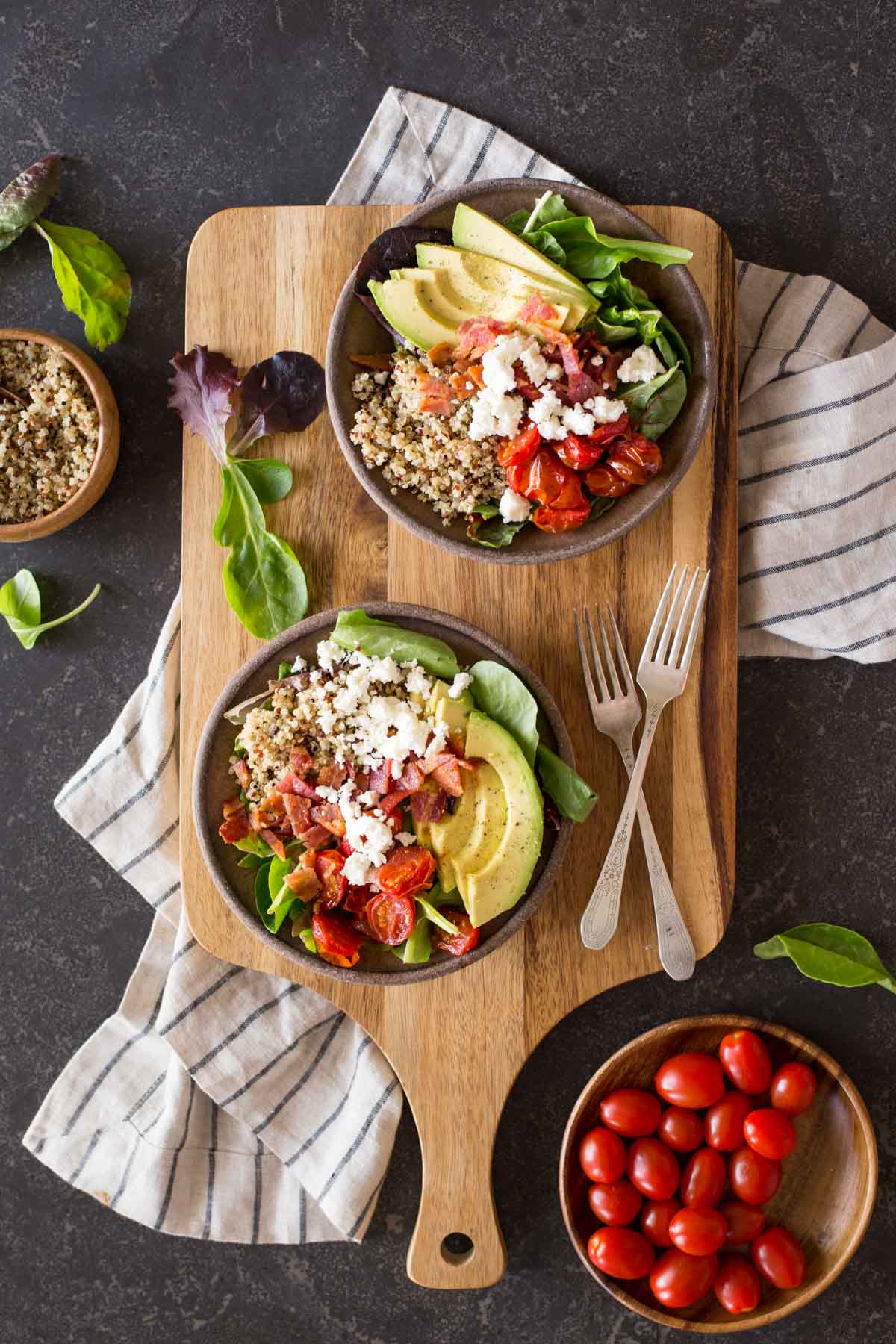 Two BLT Bliss Bowls on a cutting board with two forks, with a bowl of grape tomatoes and a bowl of quinoa next to the board. 