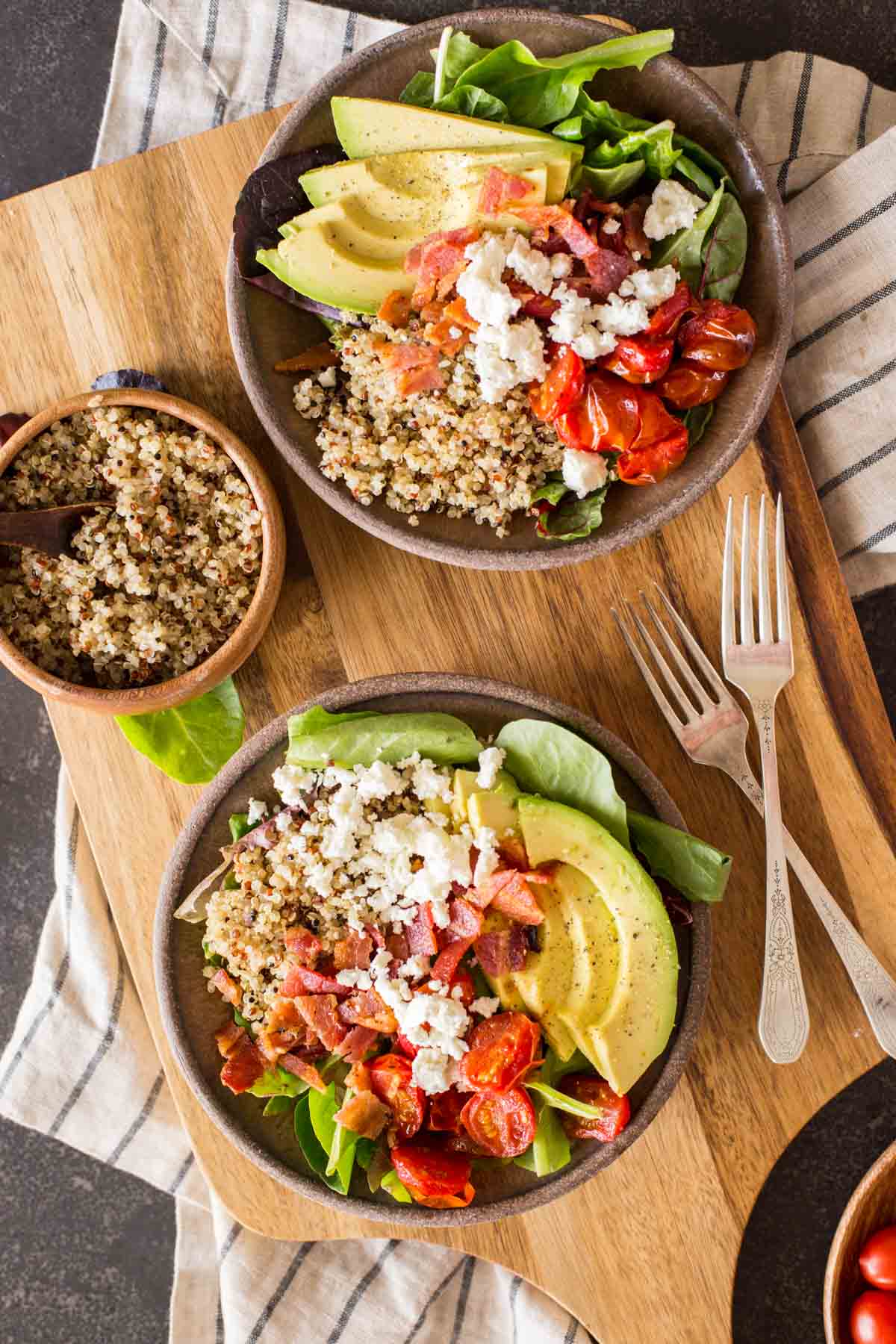 Two BLT Bliss Bowls on a cutting board with two forks, along with a bowl of quinoa.