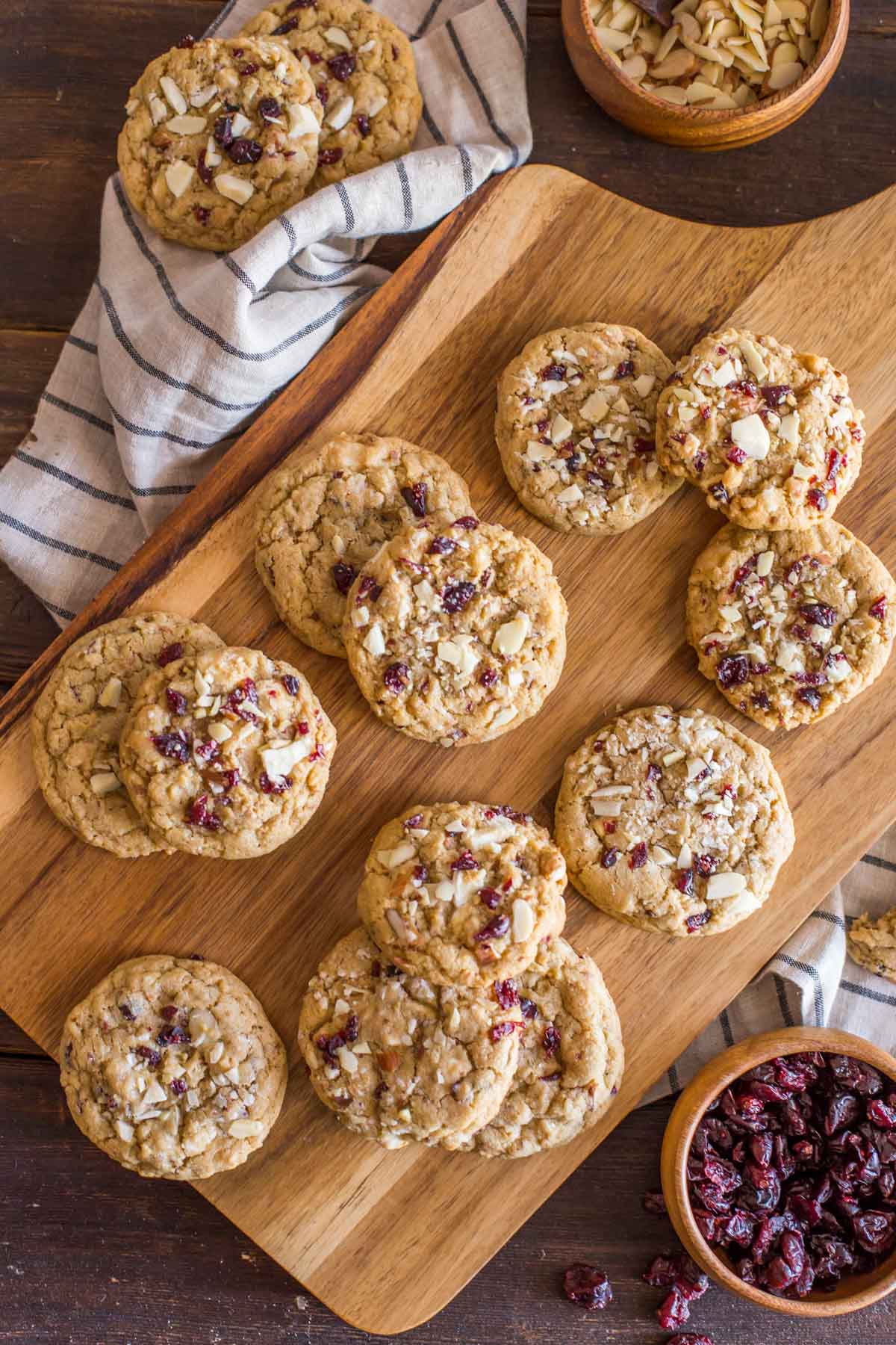Cranberry Almond White Chocolate Chunk Cookies on a wood cutting board, with a wood bowl of sliced almonds and a wood bowl of dried cranberries next to the board. 