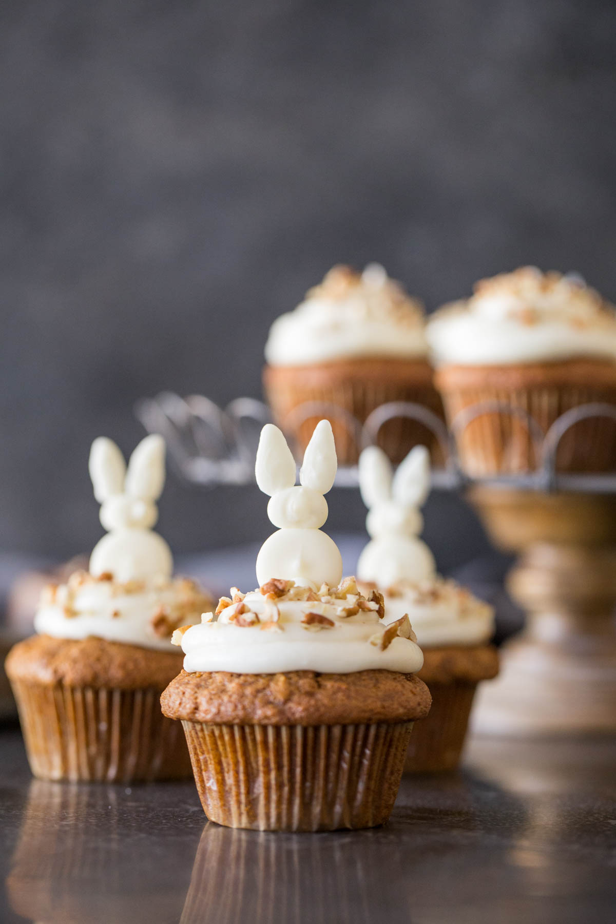 Three Carrot Cake Cupcakes with white chocolate bunnies on top, and more cupcakes on a cake stand in the background. 