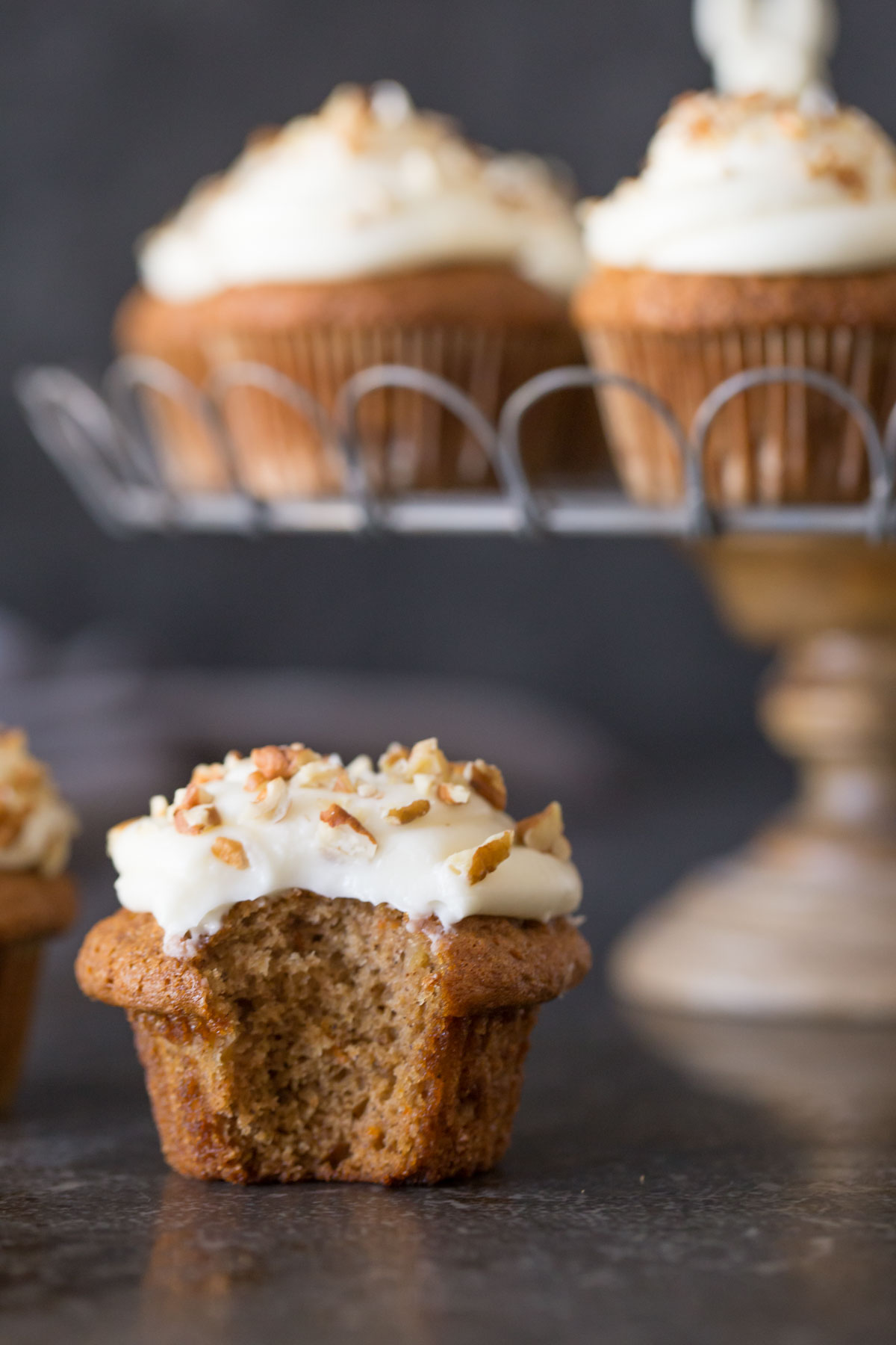 A Carrot Cake Cupcake with a bite taken out of it, with more Carrot Cake Cupcakes on a cake stand in the background. 