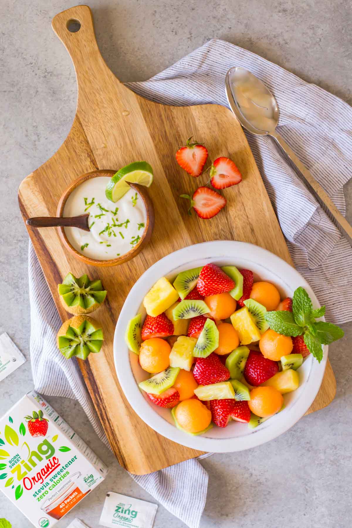 A bowl of Summer Fruit Salad sitting on a cutting board along with a small bowl of Coconut Lime Yogurt and some strawberries and kiwis, with a box of Born Sweet Zing Organic Stevia Sweetener next to the board. 