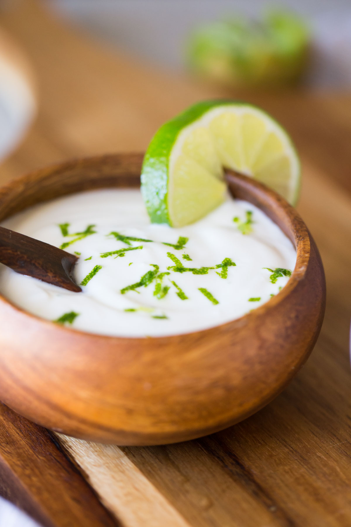 A small wood bowl of the Coconut Lime Yogurt topping, garnished with lime zest and a lime slice on the edge of the bowl. 