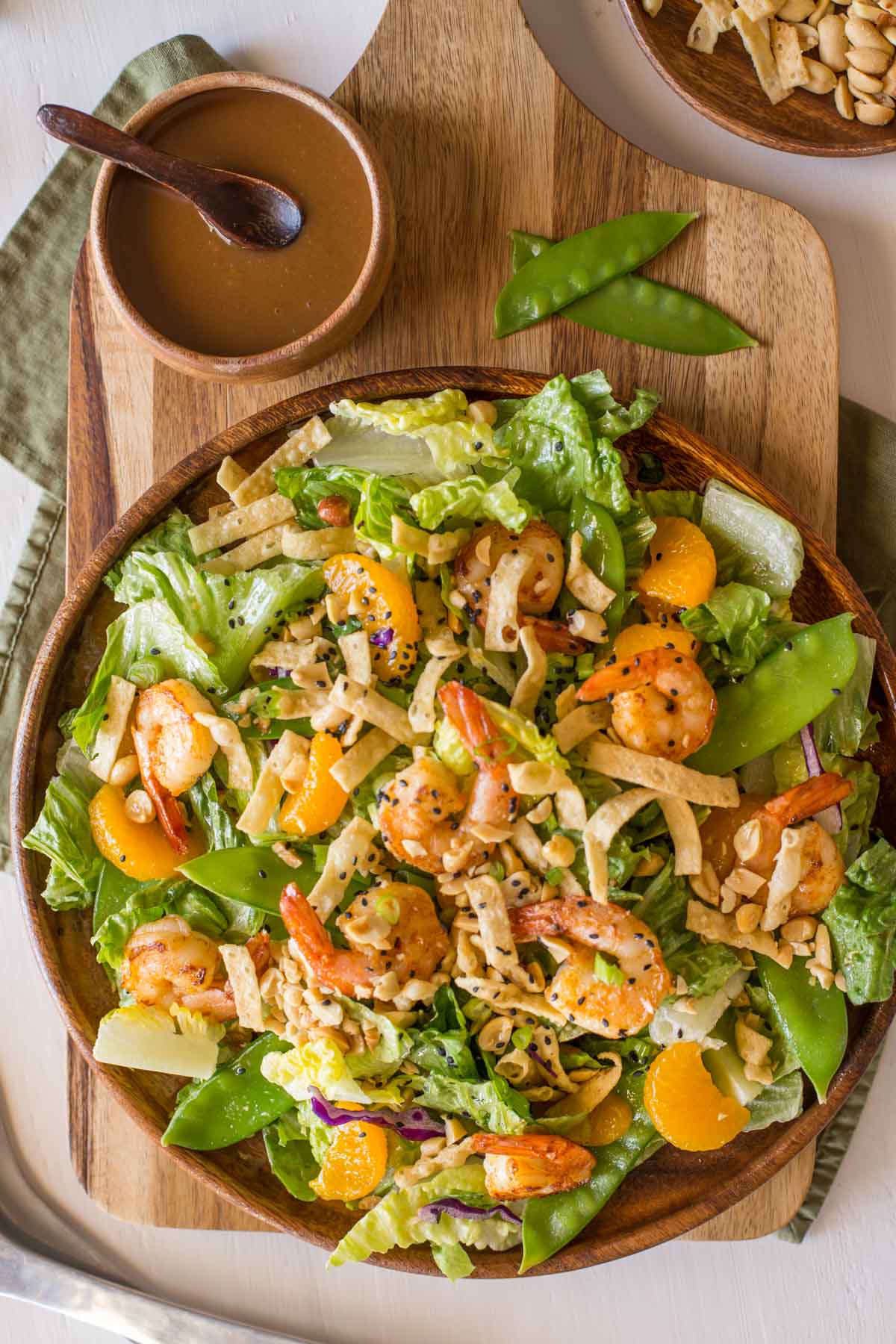 Shrimp Salad on a large serving plate, sitting next to a small wood bowl of the Spicy Peanut Dressing and a bowl of crispy wonton strips and peanuts. 