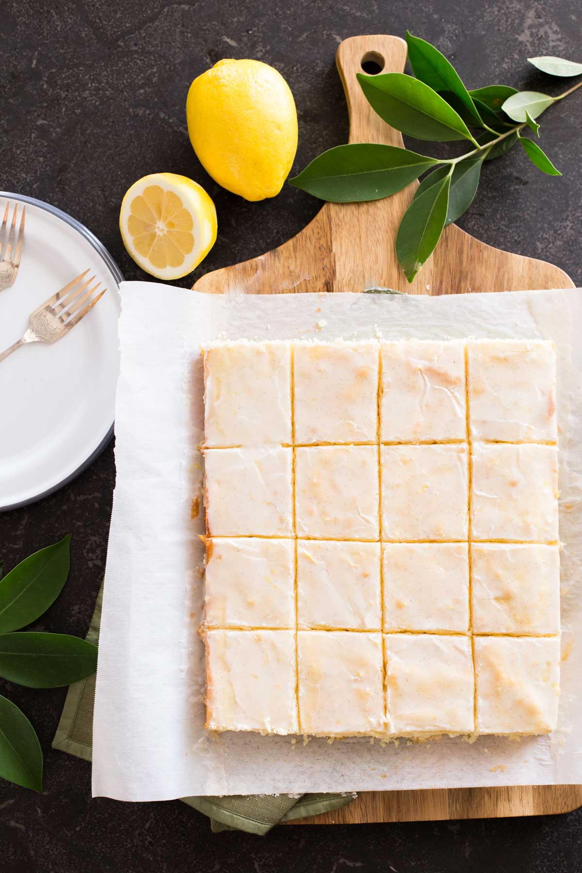Vanilla Bean Lemon Bars cut into squares on parchment paper on top of a cutting board, with lemons and tree leaves next to the board, along with some plates and forks. 