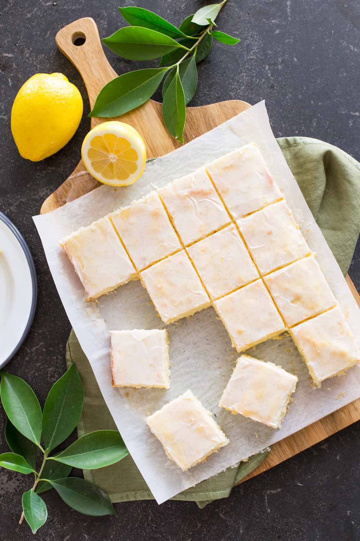 Vanilla Bean Lemon Bars cut into squares on parchment paper on top of a cutting board, with lemons and tree leaves next to the board. 