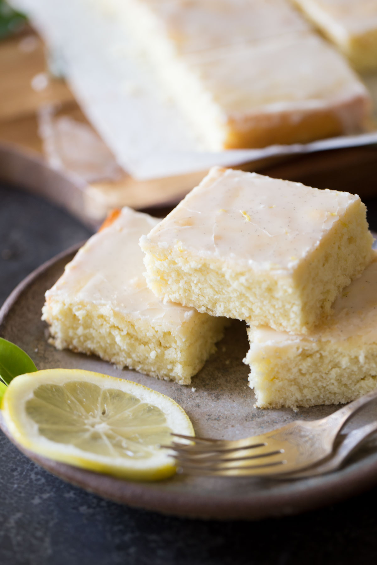Three Vanilla Bean Lemon Bars on a plate with two forks and a lemon slice, with more Vanilla Bean Lemon Bars in the background. 