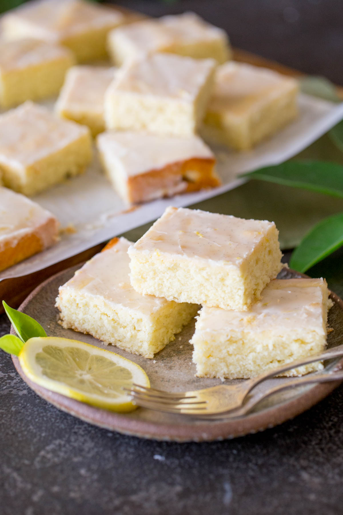 Three Vanilla Bean Lemon Bars on a plate with two forks and a lemon slice, with more Vanilla Bean Lemon Bars in the background. 