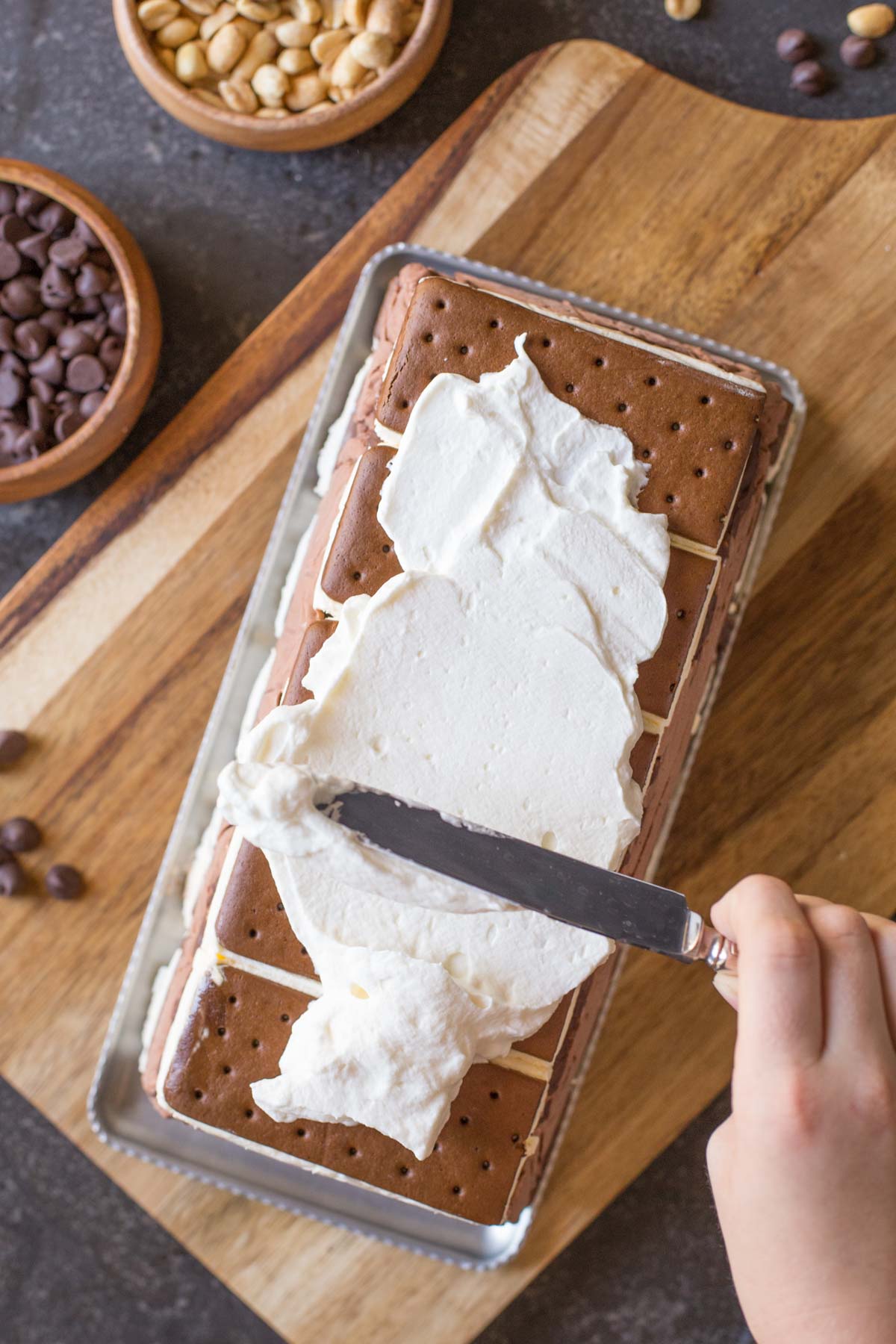 Whipped cream being spread onto the Chocolate Peanut Butter Ice Cream Slice Cake, that is sitting on a platter on top of cutting board, with a bowl of chocolate chips and a bowl of peanuts next to it. 