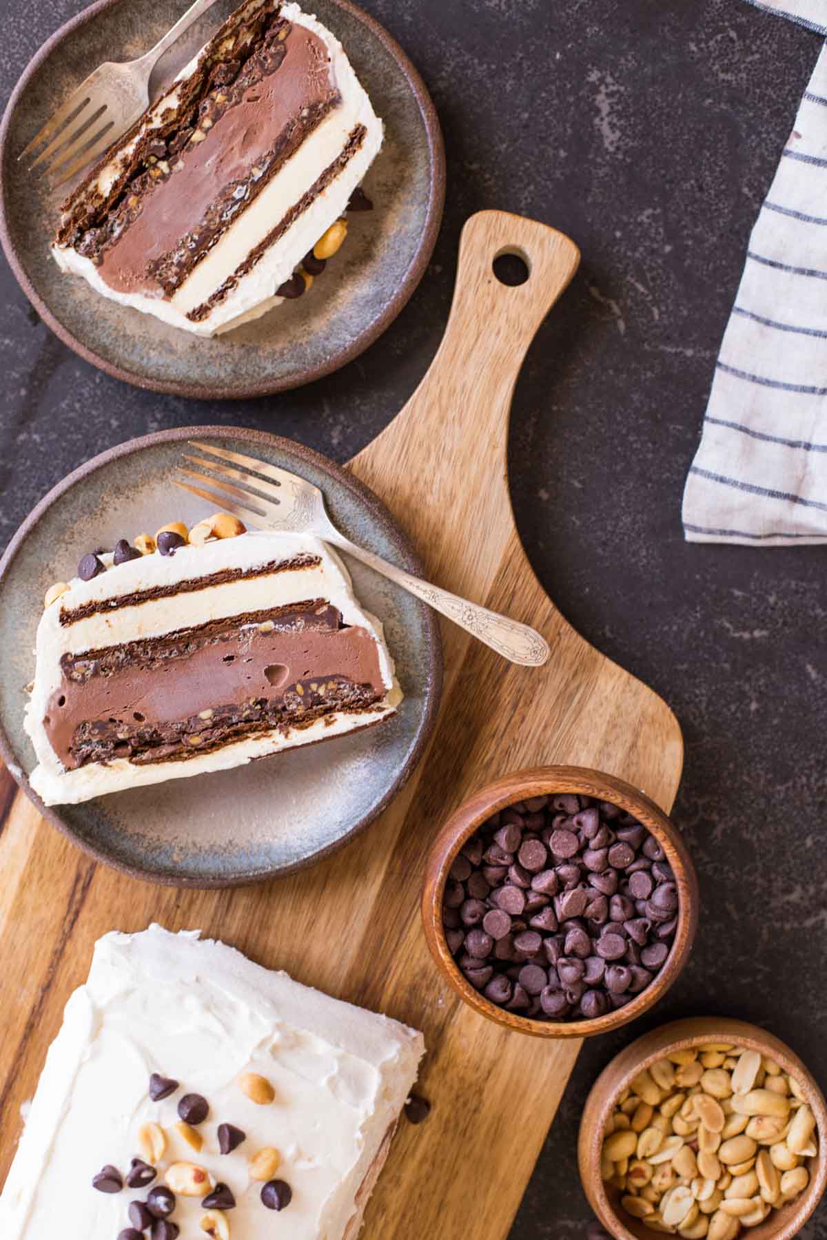 Two slices of Chocolate Peanut Butter Ice Cream Slice Cake on plates with forks, sitting next to the rest of the cake, along with a bowl of chocolate chips and a bowl of peanuts. 