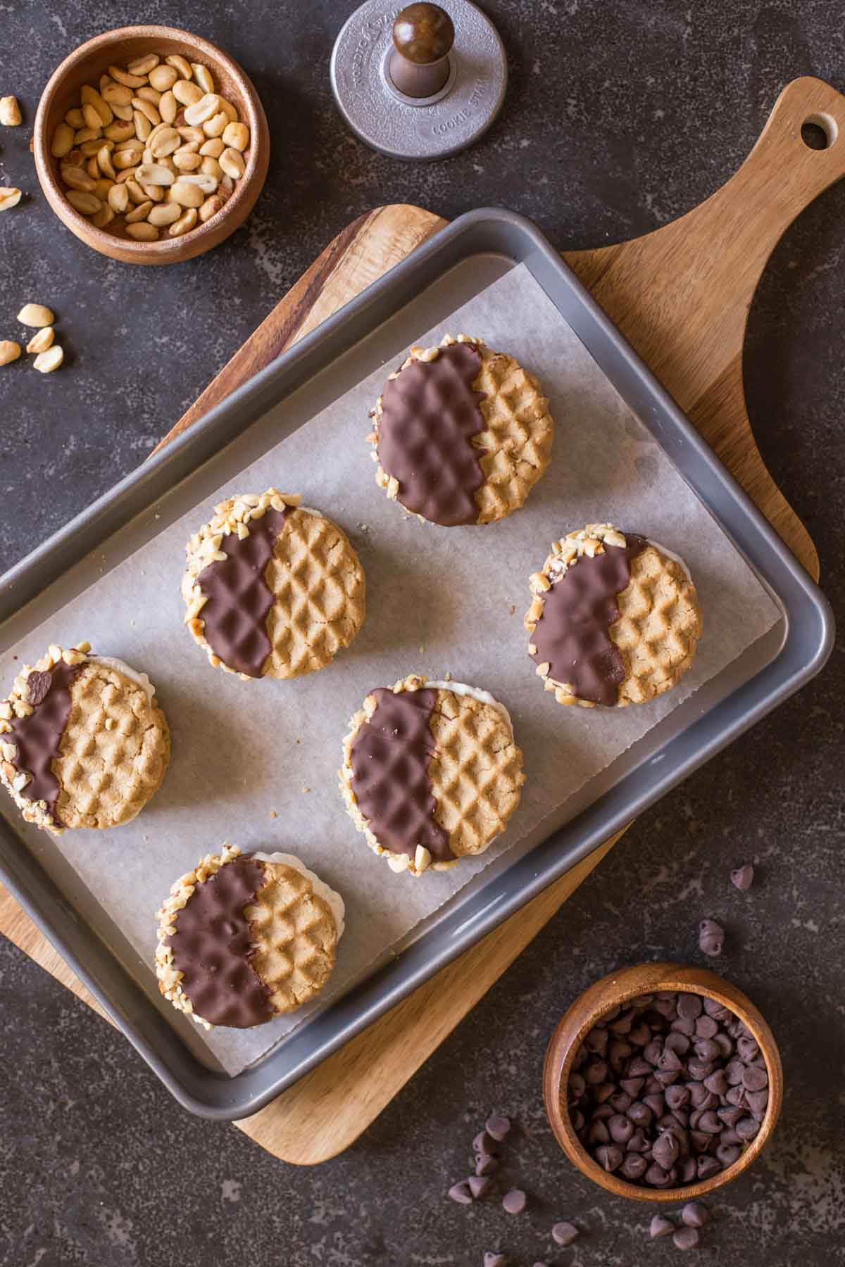 Peanut Butter Cookie Ice Cream Sandwiches on a parchment paper lined baking sheet, with a bowl of peanuts and a bowl of chocolate chips next to it. 