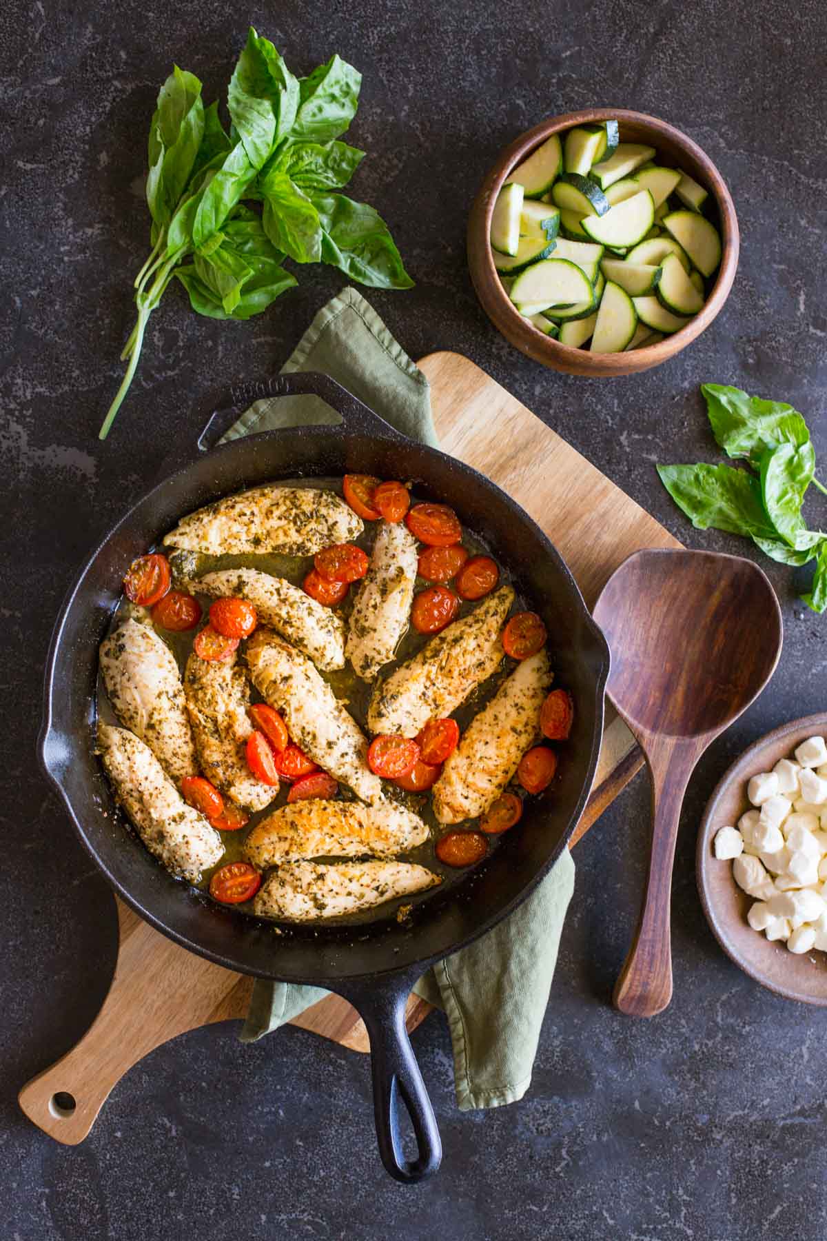 Cooked chicken and caramelized cherry tomatoes in a cast iron skillet, with some fresh basil, a bowl of sliced zucchini, a bowl of fresh mozzarella pearls and a wooden spoon next to the skillet, all to make the Pesto Chicken Skillet.