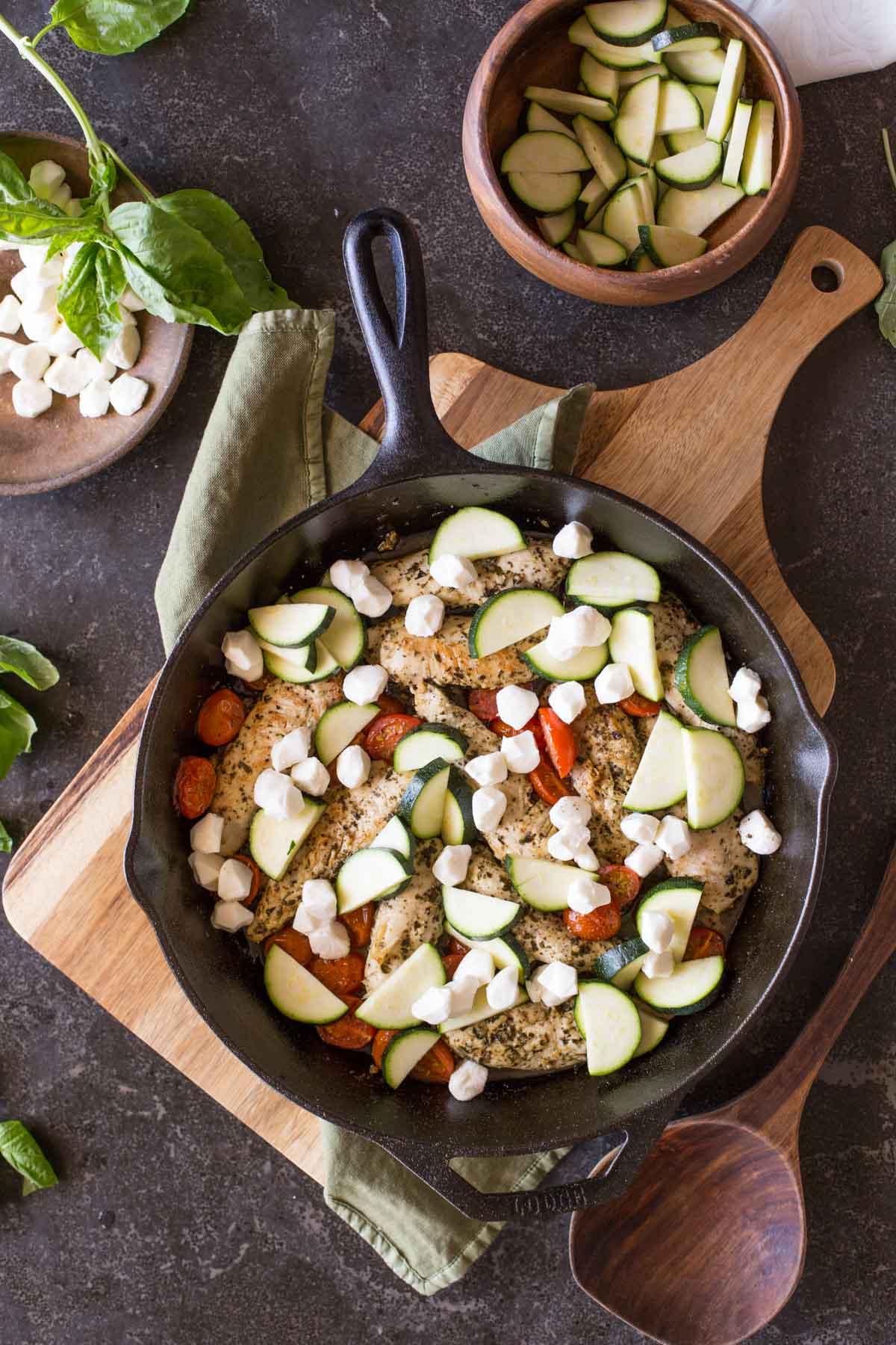 Cooked chicken and caramelized cherry tomatoes in a cast iron skillet, topped with zucchini slices and fresh mozzarella pearls, with some fresh basil, a bowl of sliced zucchini, a bowl of fresh mozzarella pearls and a wooden spoon next to the skillet.
