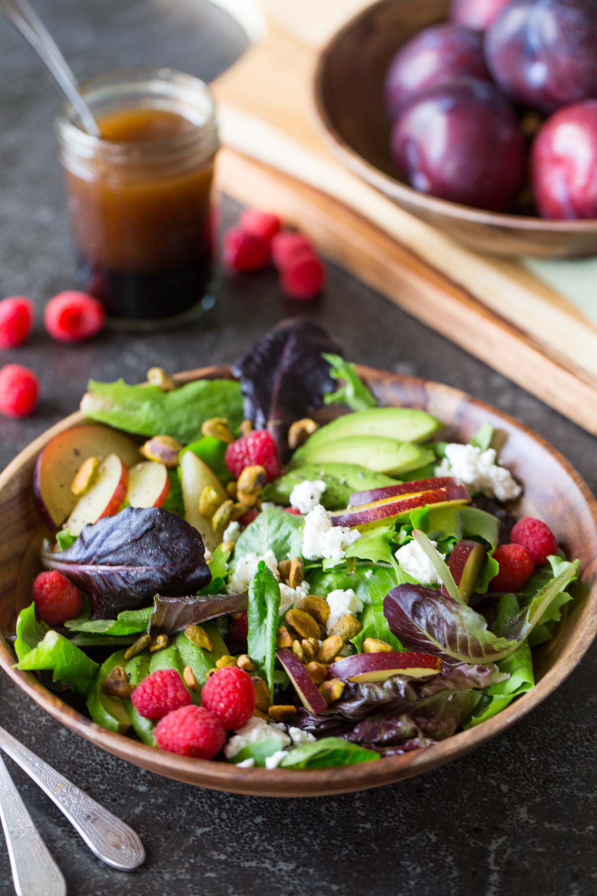 Summer Fruited Salad with Goat Cheese and Pistachios in a wood bowl, with a jar of homemade balsamic vinaigrette in the background along with a bowl of whole plums.  