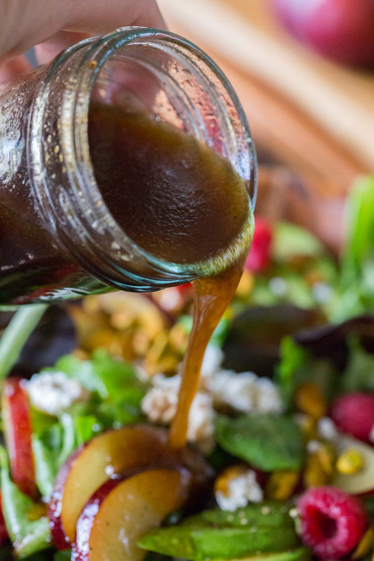 A glass jar of homemade balsamic vinaigrette being poured on top of the Summer Fruited Salad with Goat Cheese and Pistachios.