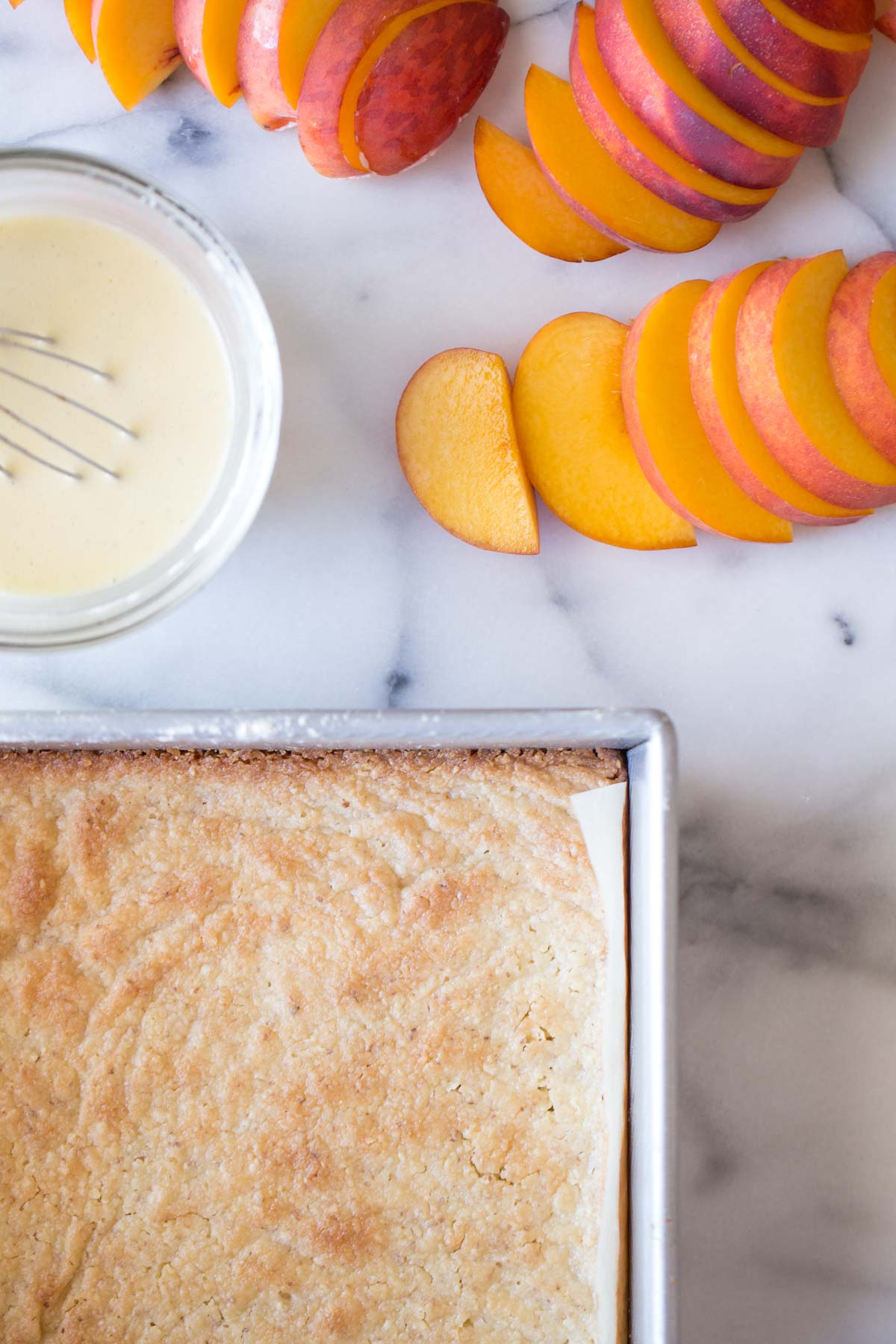 Almond shortbread crust in a baking dish, with some sliced fresh peaches and a bowl of creamy vanilla custard sitting next to it.  