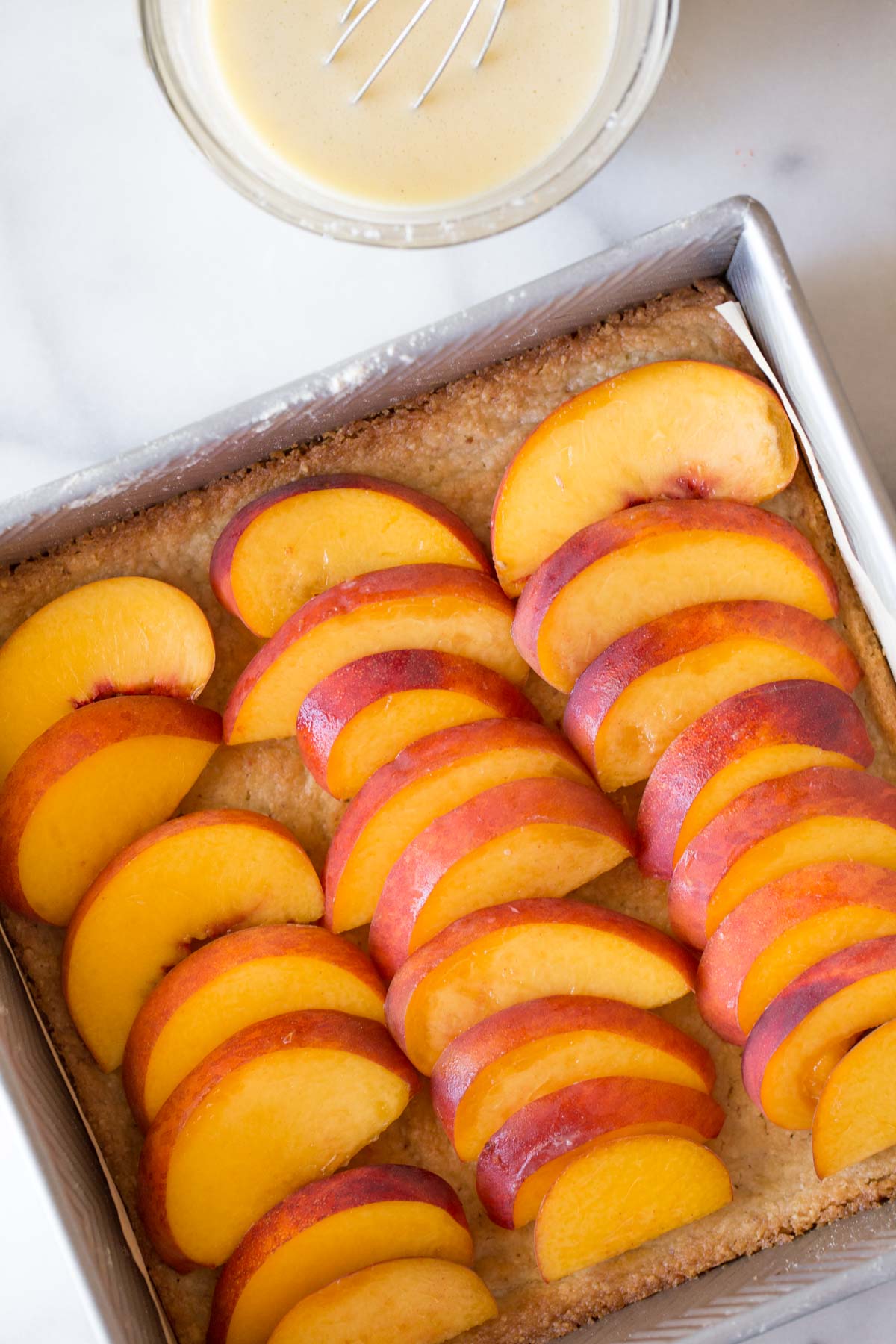 A baking dish with the fresh sliced peaches placed on top of the almond shortbread crust, with a bowl of creamy vanilla custard next to it. 