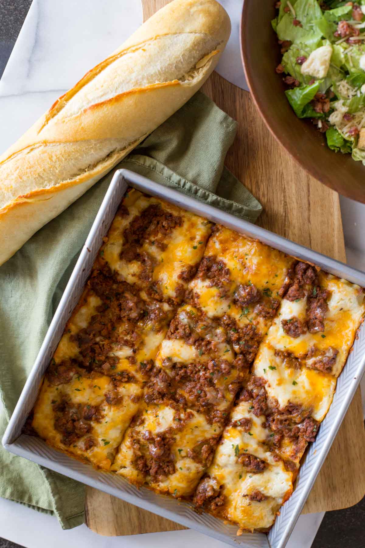 Simple Homemade Lasagna in a square baking dish, with a loaf of French bread and a bowl of salad next to it. 
