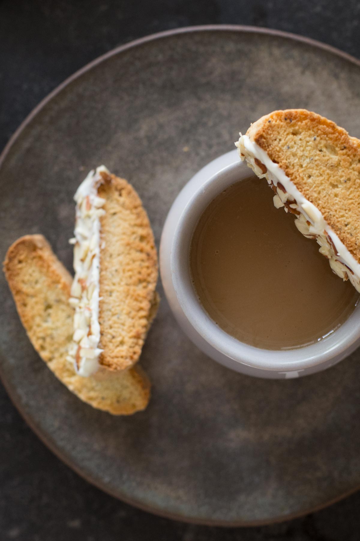 Two Almond Poppy Seed Biscotti slices on a plate, along with a cup of coffee with an Almond Poppy Seed Biscotti slice balancing on the rim of the cup.  