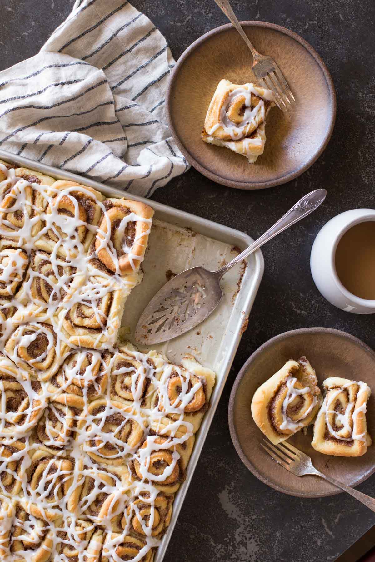 Sheet Pan Cinnamon Rolls on two plates with forks, sitting next to a cup of coffee and the baking sheet of the rest of the cinnamon rolls with a serving utensil.  