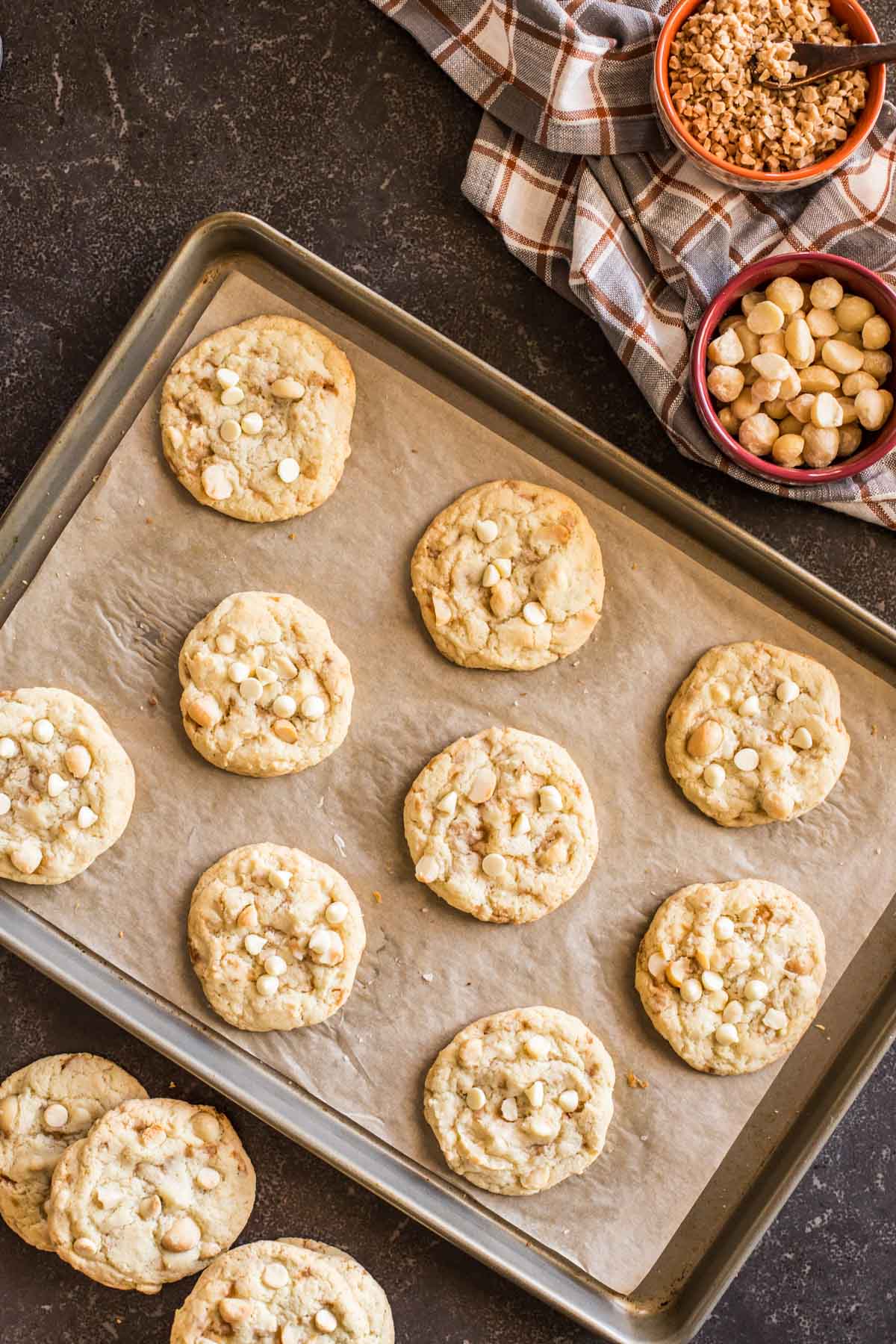 Buttery Toffee White Chocolate Chip Macadamia Nut Cookies on a parchment paper lined baking sheet, with more cookies sitting next to the pan, as well as a bowl of roasted salted macadamia nuts and a bowl of toffee bits.