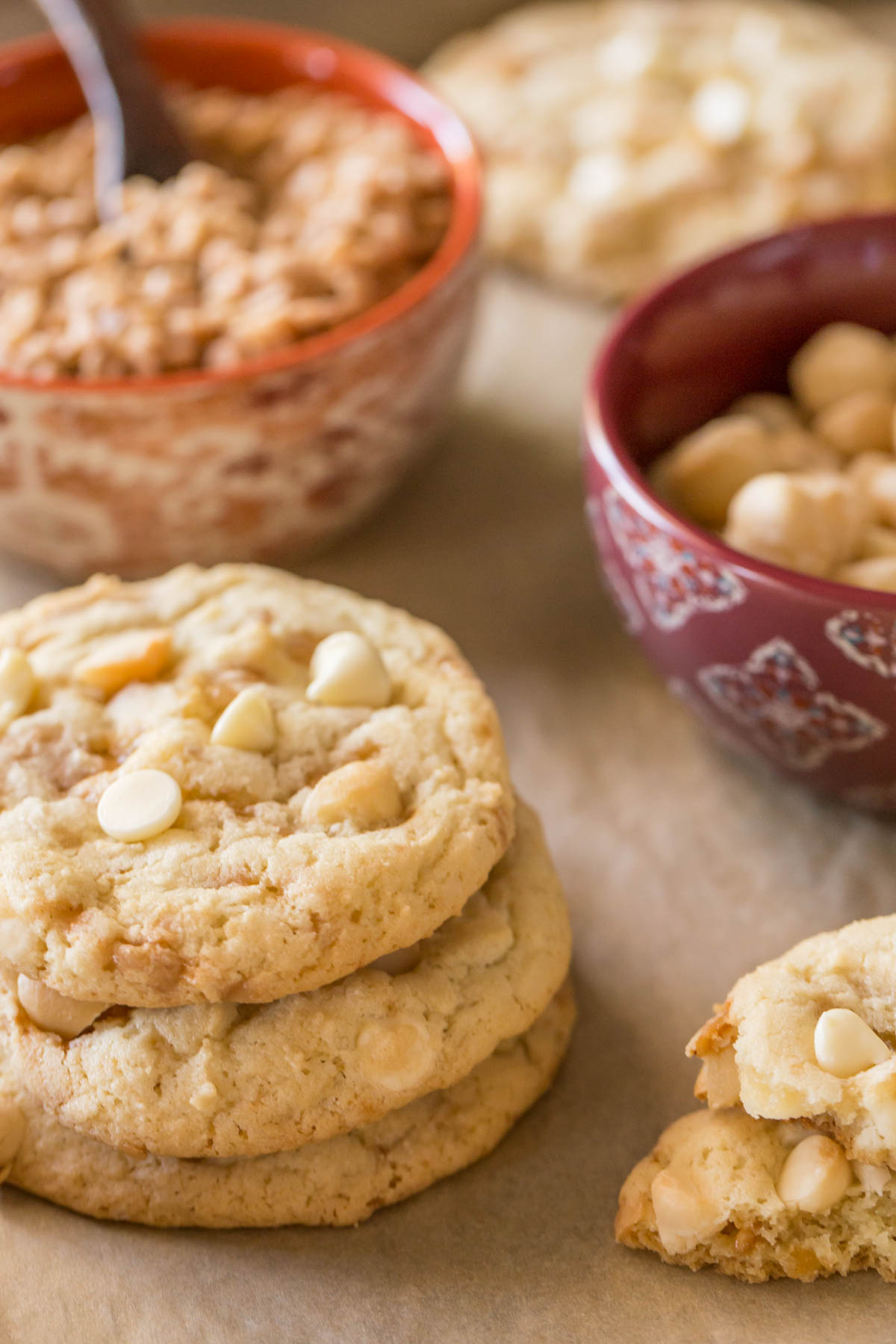 Three Buttery Toffee White Chocolate Chip Macadamia Nut Cookies stacked on top of each other, with a bowl of roasted salted macadamia nuts and a bowl of toffee bits in the background. 