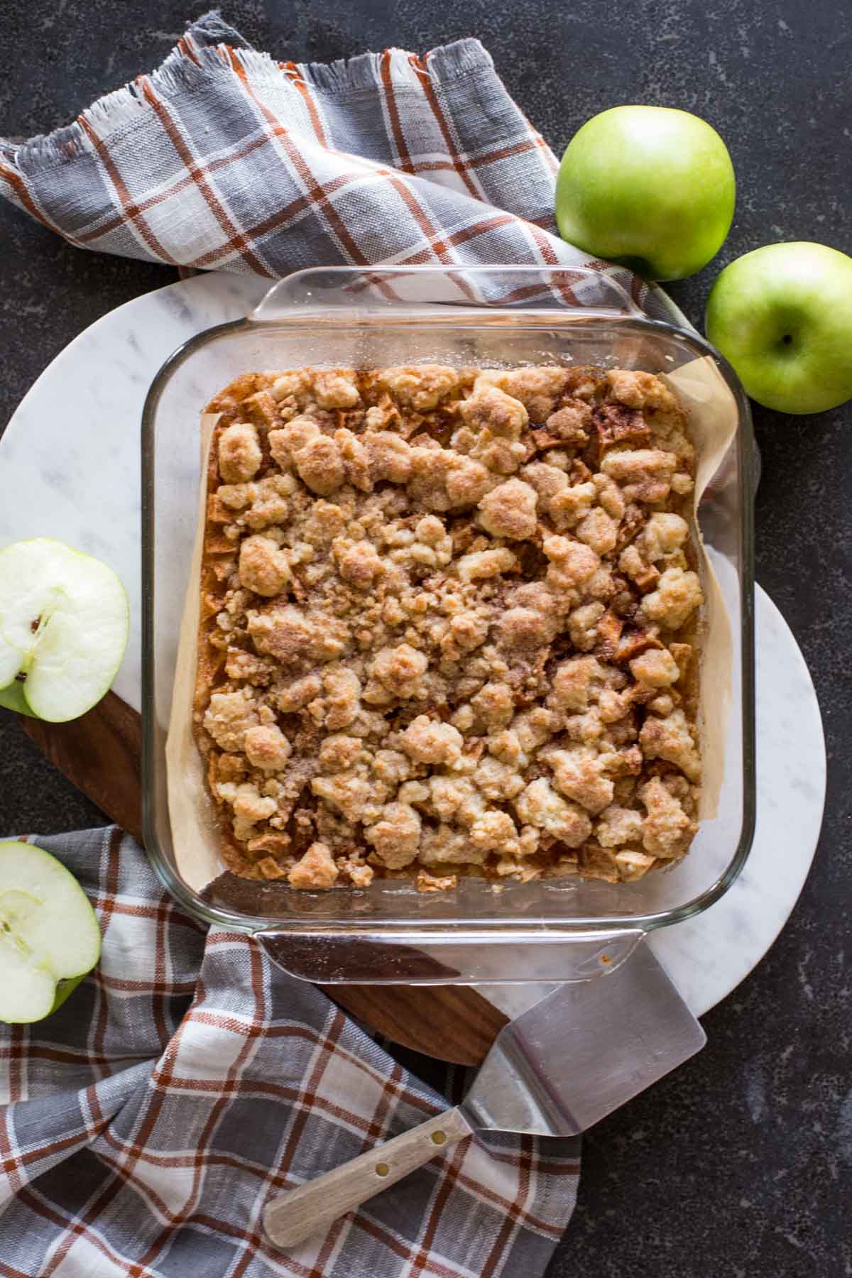 Apple Pie Crumb Bars in a square baking dish, with whole and halved apples next to it, as well as a serving spatula. 