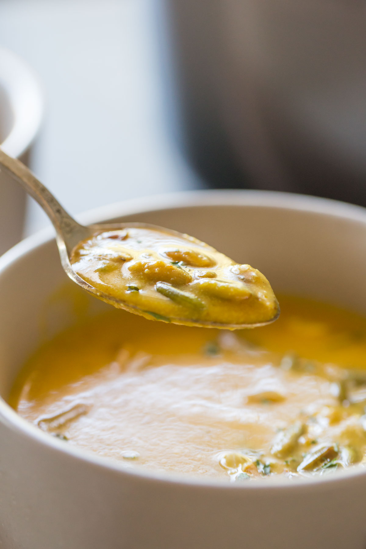 A bowl of Creamy Cashew Pumpkin Soup, garnished with pepitas, chopped cashews and parsley, with a spoon full of soup over the bowl. 