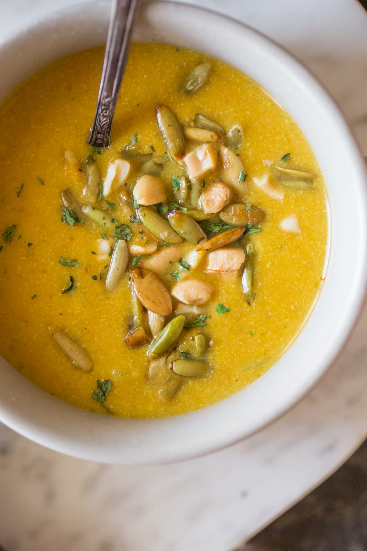A bowl of Creamy Cashew Pumpkin Soup, garnished with pepitas, chopped cashews and parsley, with a spoon in it. 