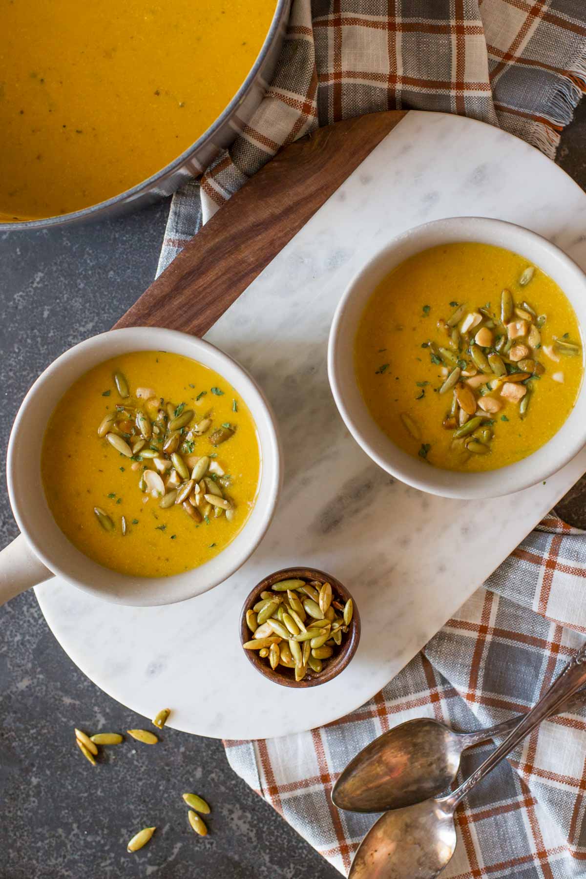 Two bowls of Creamy Cashew Pumpkin Soup, garnished with pepitas, chopped cashews and parsley, sitting next to the pot of soup, a small bowl of pepitas and two spoons. 