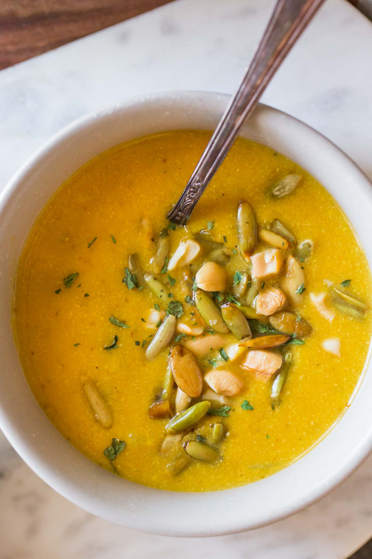 A bowl of Creamy Cashew Pumpkin Soup, garnished with pepitas, chopped cashews and parsley, with a spoon in it. 