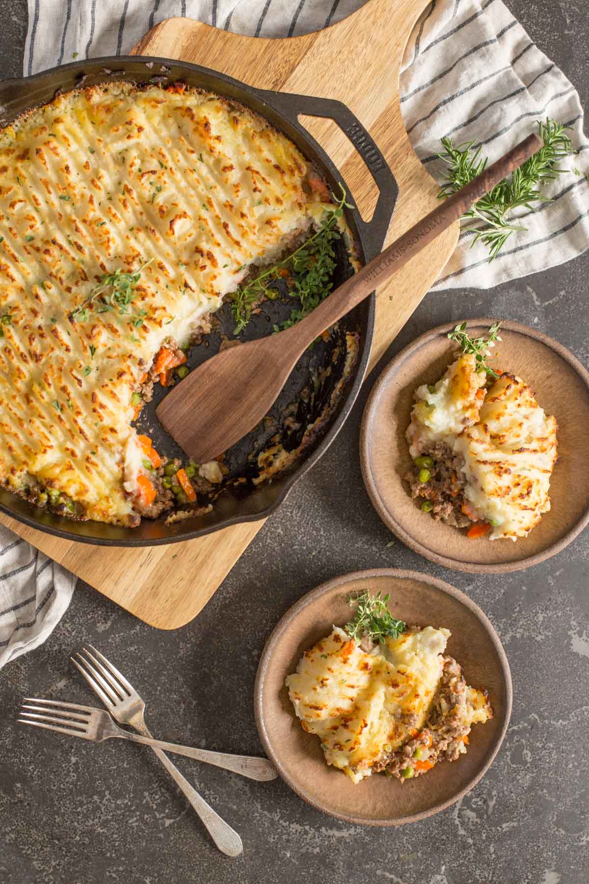 Two servings of Easy Homemade Shepherd's Pie dished on to two plates, with two forks next to them as well as the cast iron skillet of Easy Homemade Shepherd's Pie with a wooden spoon in it. 