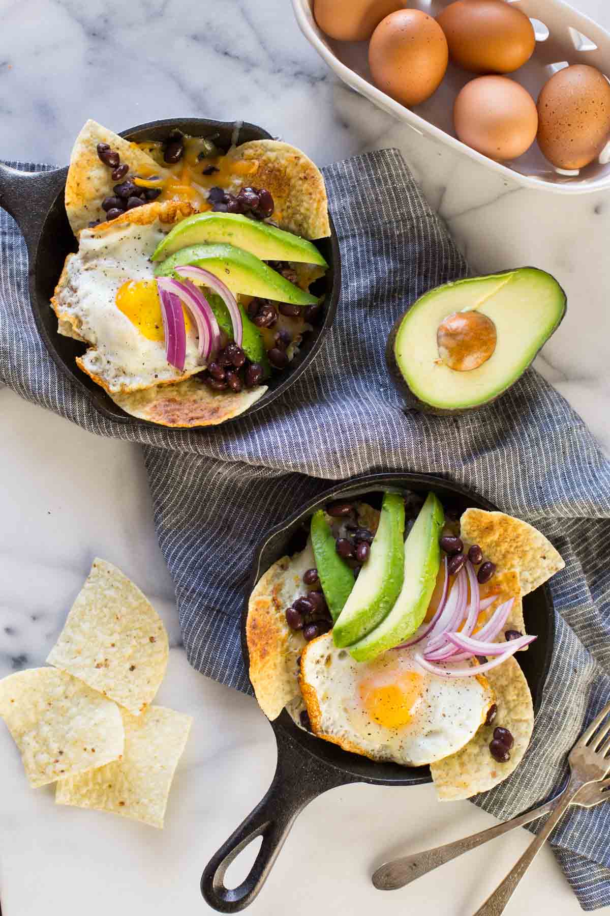 Two Breakfast Nacho Skillets topped with avocado slices and red onions, with a few tortilla chips, a half of an avocado, a basket of eggs and two forks sitting next to the skillets. 