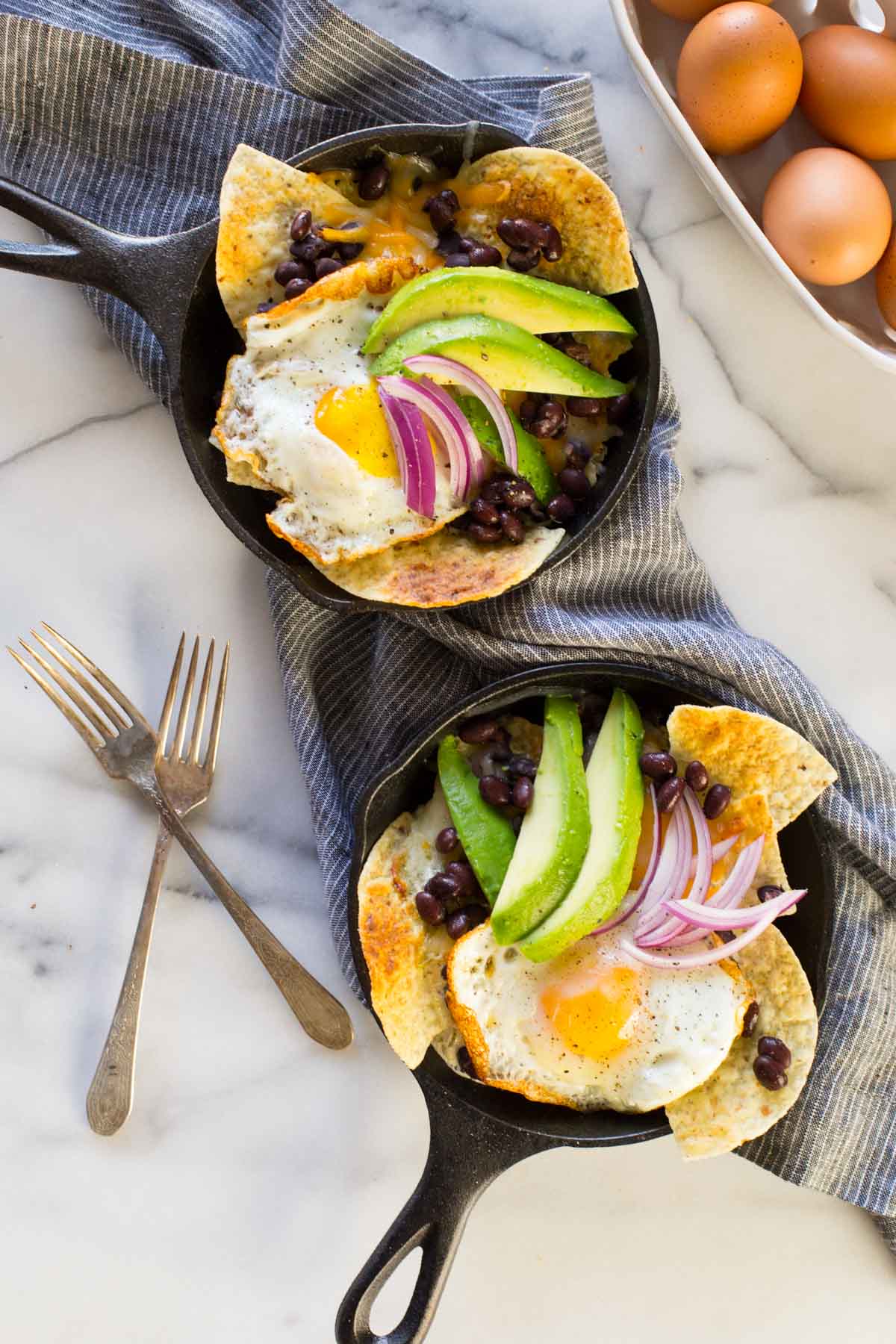 Two Breakfast Nacho Skillets topped with avocado slices and red onions, with two forks and a basket of eggs sitting next to the skillets. 