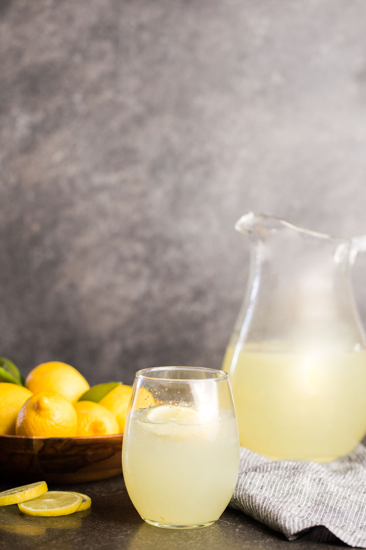 Lemonade in glass pitcher with bowl of lemons and glass of lemonade. 