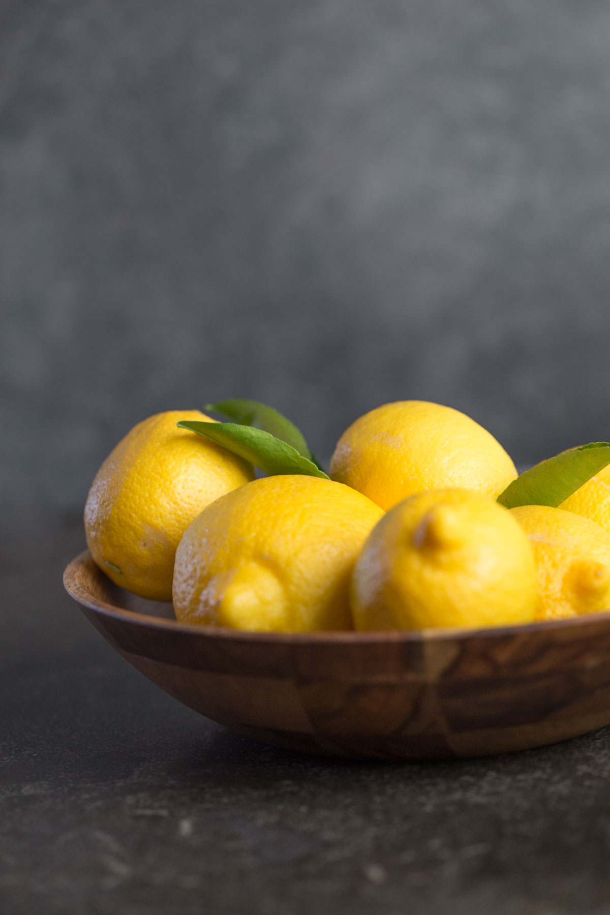 Wooden bowl of lemons with leaves attached from the front view with a dark grey background. 