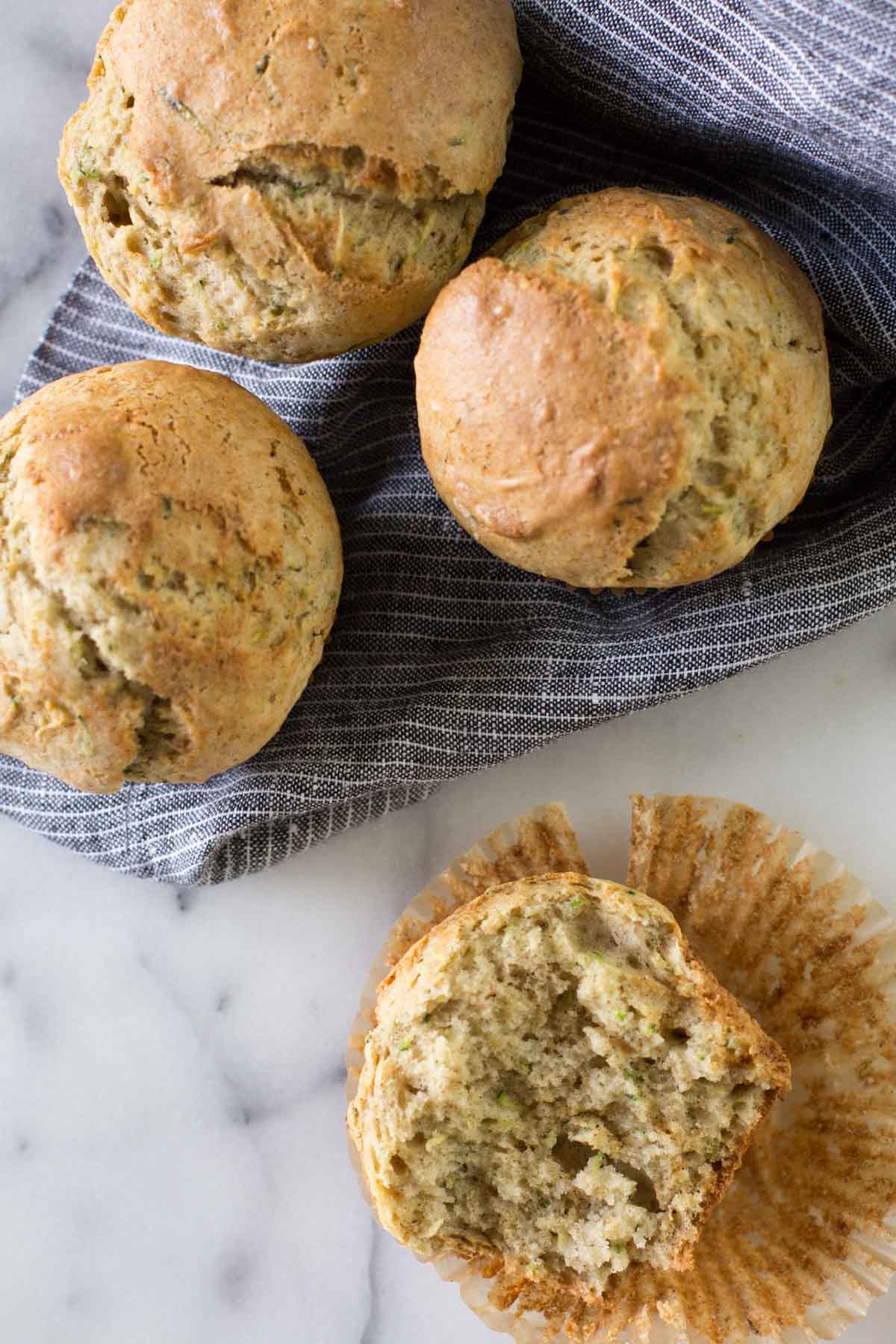 A half of a Healthier Zucchini Muffin sitting on its wrapper, with three more whole muffins sitting on a cloth napkin. 