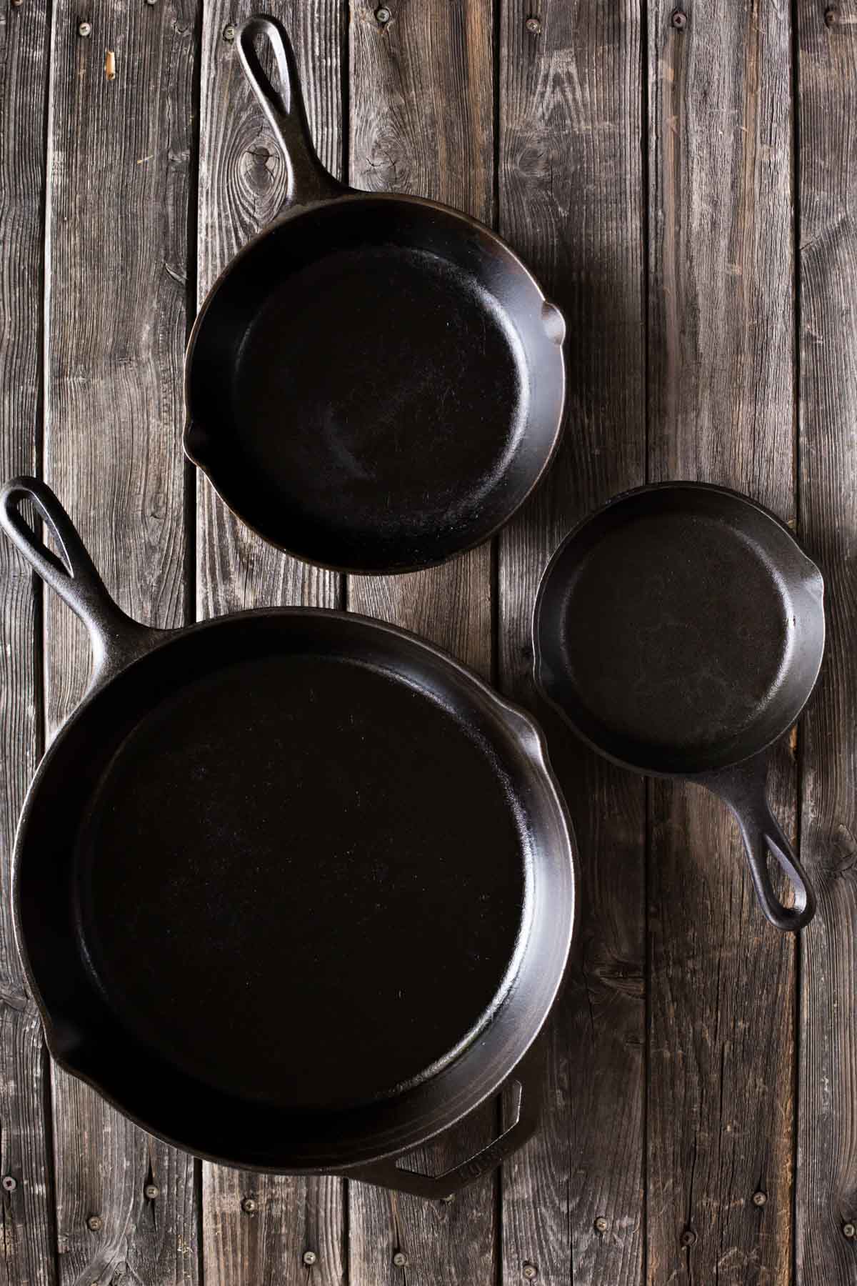 A top down view of a small, medium, and large sized cast iron skillet on a wooden background. 