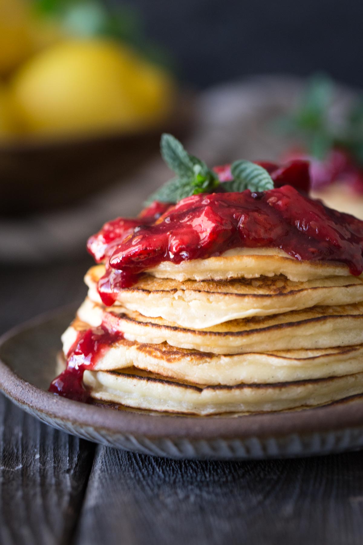 A close up view of a stack of lemon ricotta pancakes and fresh berry sauce on a plate with lemons in the background. 