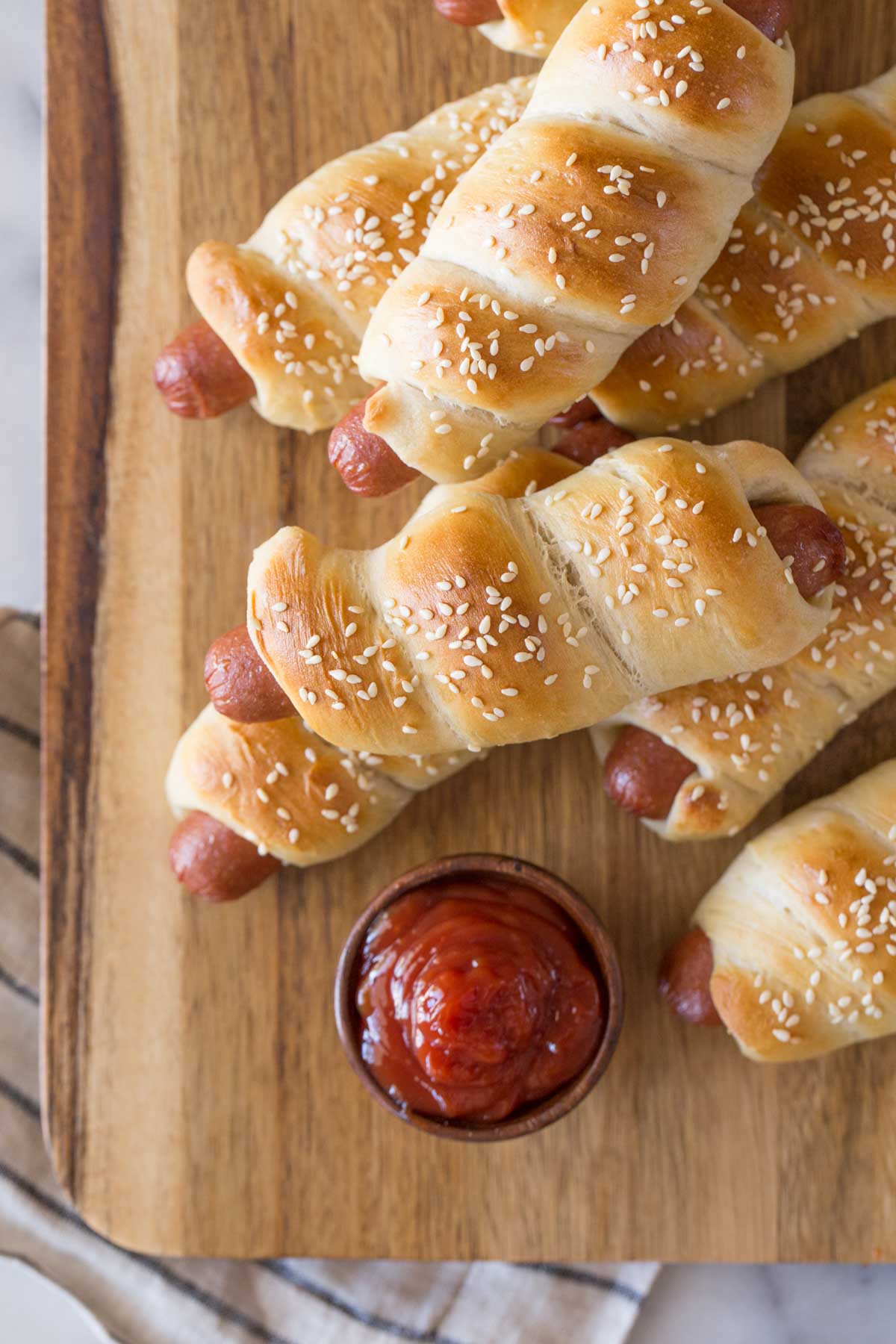Homemade Pigs in a Blanket on a wooden cutting board with a bowl of ketchup. 