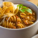 A close up shot of a grey bowl filled with Beef Chili Queso with pasta, Fritos, source cream, and cilantro.